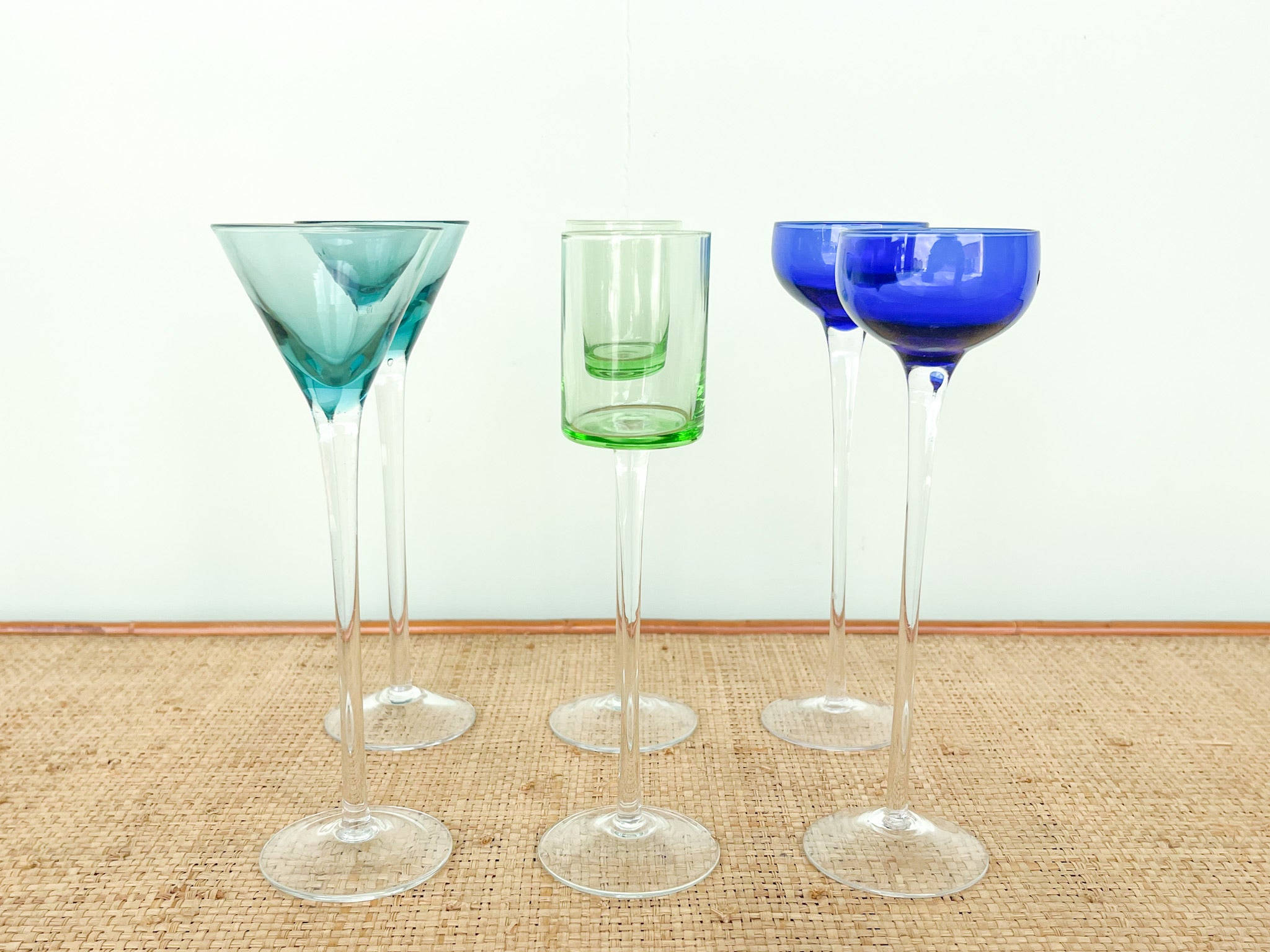 「優雅糖漿杯於高雅場景」 壁紙 — 四個復古雞尾酒杯，帶有青綠色和鈷藍色的彩色杯身，放置在編織黃麻表面上，背景是純白色牆壁。