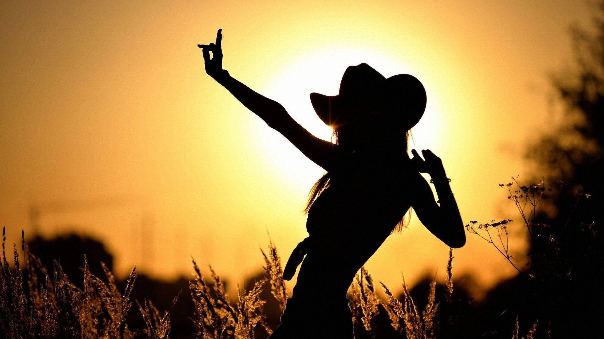 優雅舞動的牛仔女 壁紙 — 一個戴著牛仔帽的人影，在燦爛的夕陽天空下歡快地跳舞。