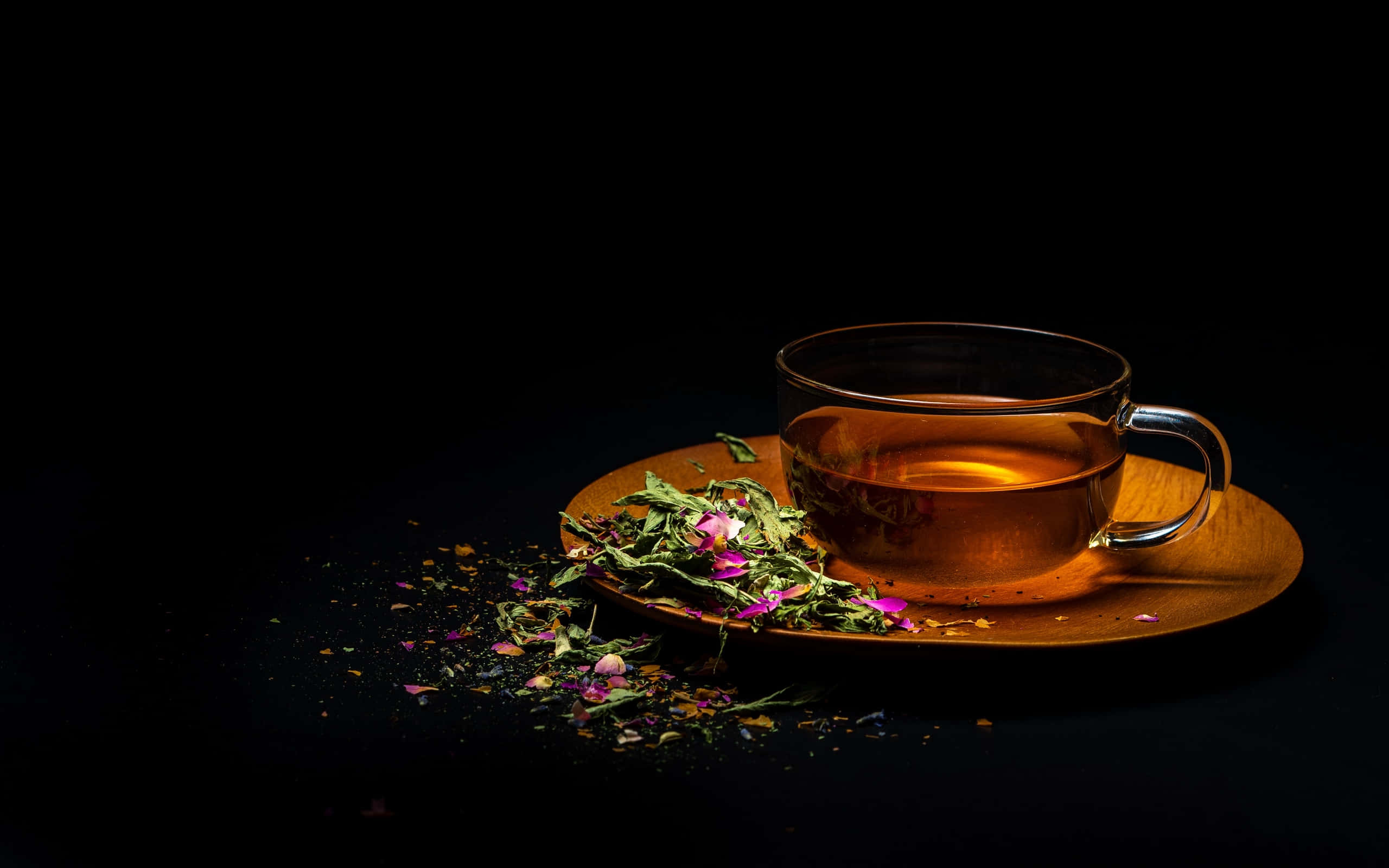 優雅設計茶杯 壁紙 — 一個裝滿琥珀色茶的玻璃茶杯放在木製茶托上，周圍散落著乾燥的玫瑰花瓣和葉子，背景為黑色。