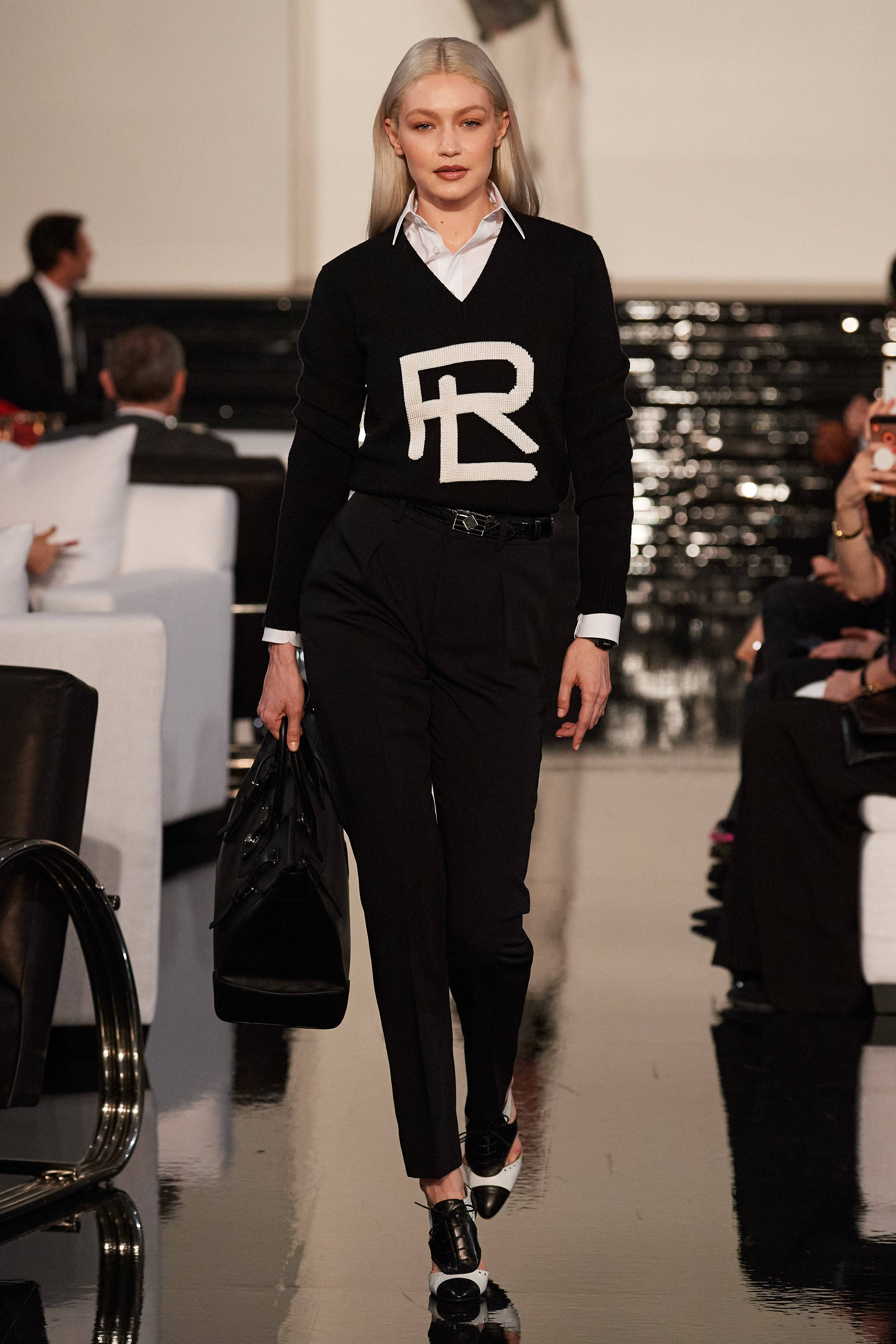 優雅象徵 - Ralph Lauren 黑色標誌 壁紙 — 一位模特兒自信地走在光亮的時裝天橋上，身穿帶有白色標誌的黑色毛衣，搭配黑色長褲和高跟鞋。