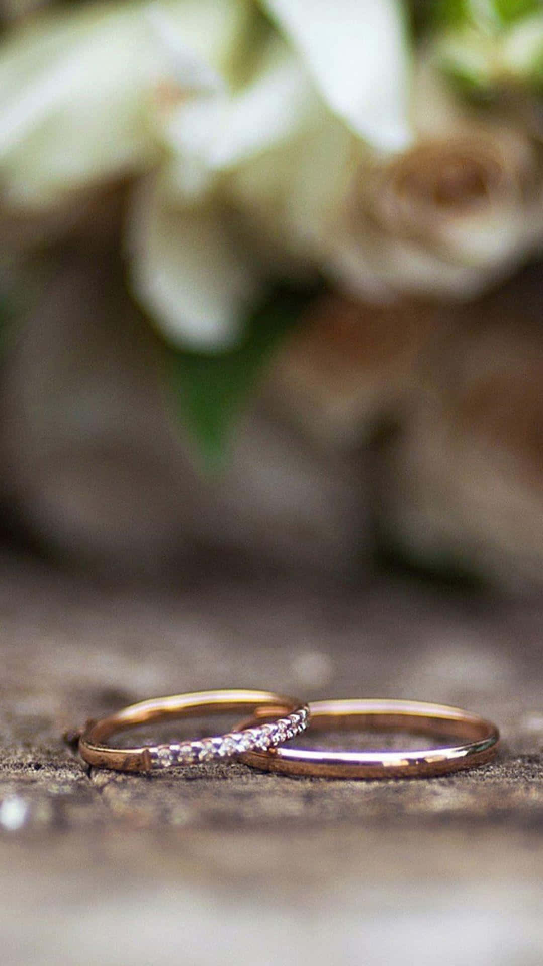 優雅鑽石戒指特寫 壁紙 — 兩枚玫瑰金結婚戒指放在風化木上，背景有柔和的白色花朵模糊。