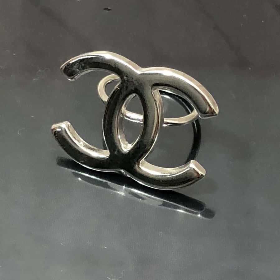優雅香奈兒標誌 壁紙 — 一枚時尚的銀色 Chanel 標誌戒指在深色反光表面上閃耀，帶有銳利的高光和柔和的陰影。