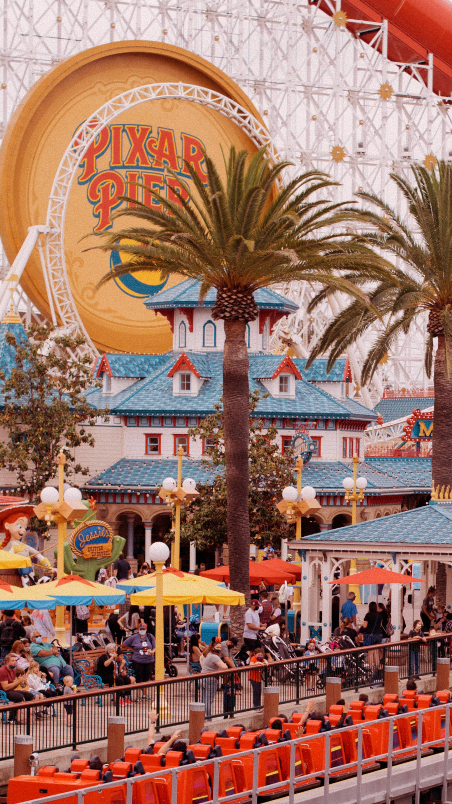 迪士尼 桌布 壁紙 — 充滿活力的 Pixar Pier 主題公園場景，有棕櫚樹、彩色遮陽傘和一個巨大的 Pixar 標誌，在明亮的日光下。