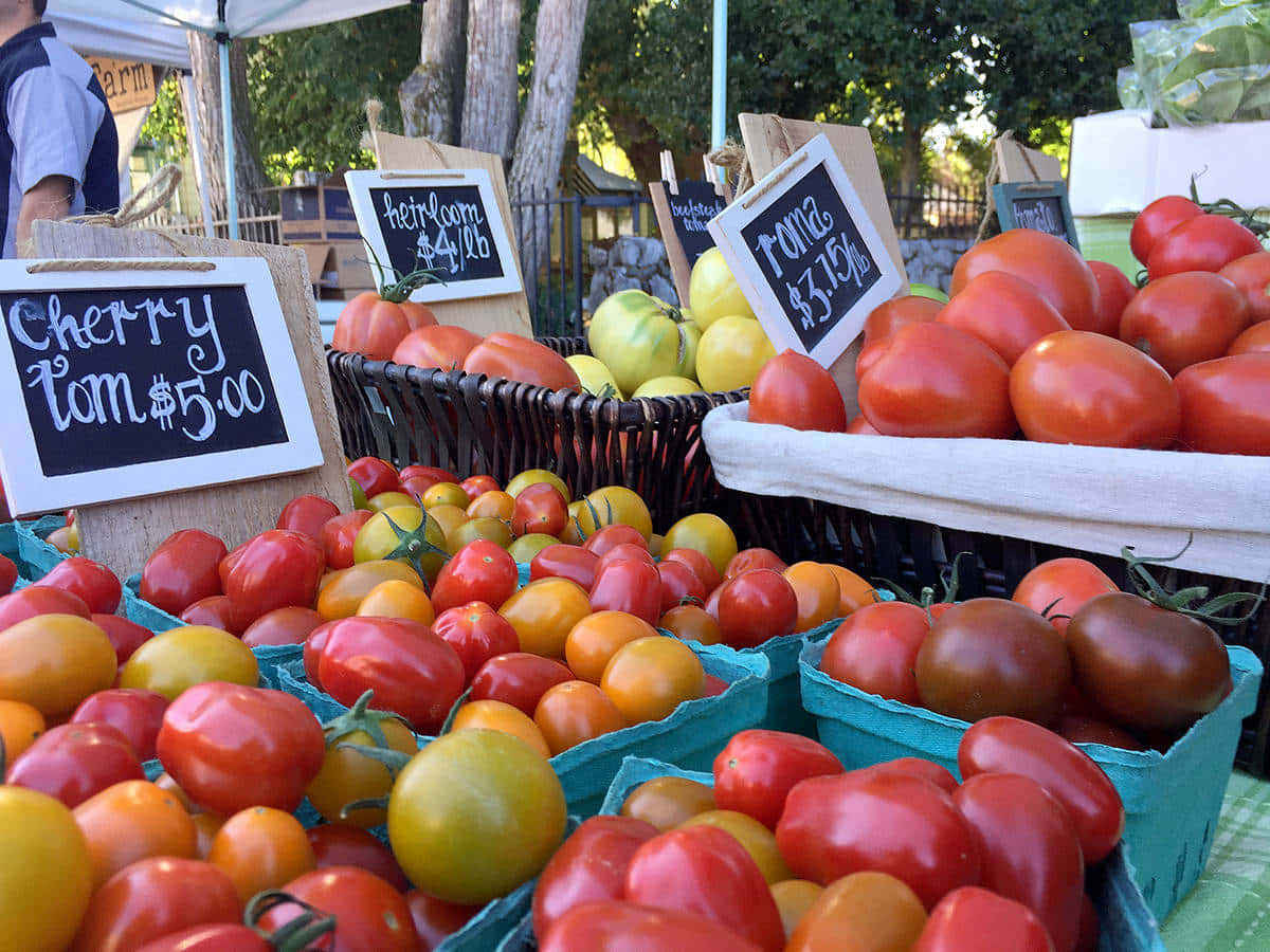 Farmers Market 桌布 壁紙 — 充滿活力的傳家寶番茄籃子，呈現紅色、黃色和橙色色調，在陽光明媚的戶外農貿市場攤位上展示。
