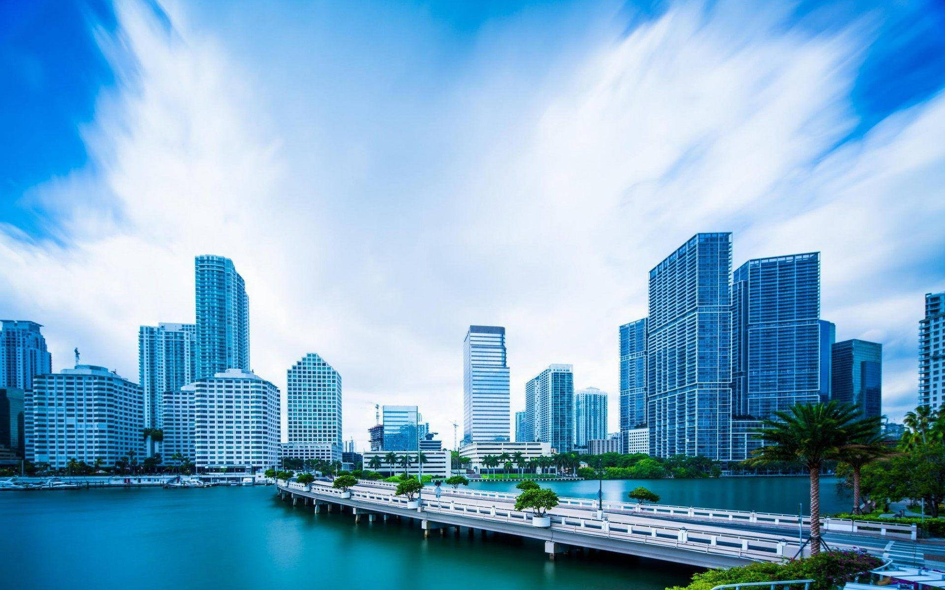 View 桌布 壁紙 — 充滿活力的城市天際線，現代摩天大樓倒映在碧綠色的水中，動態的藍天上有條紋狀的雲彩。