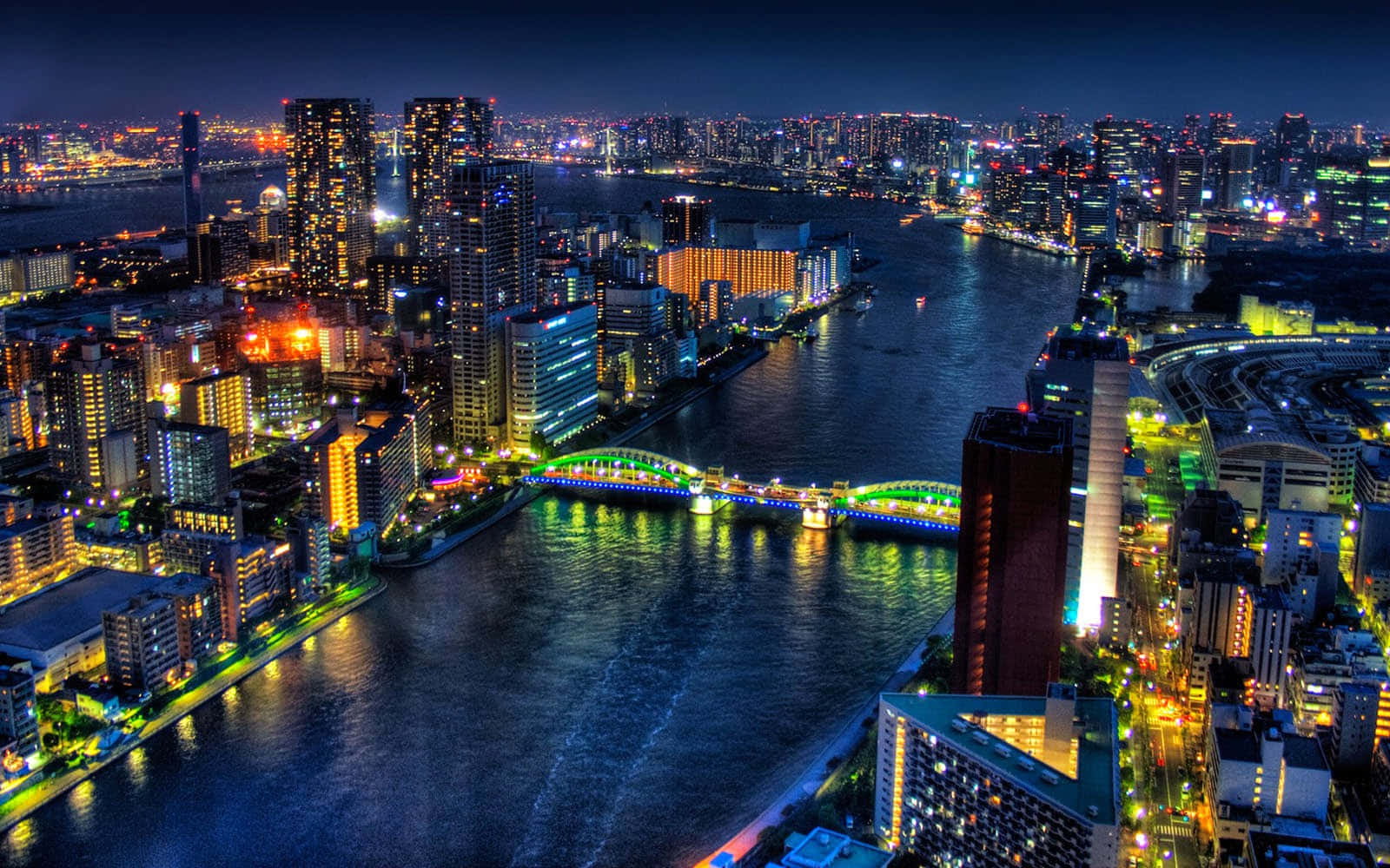 東京 桌布 壁紙 — 充滿活力的城市景觀夜間鳥瞰圖，閃爍的摩天大樓和橫跨黑暗河流的彩色橋樑。
