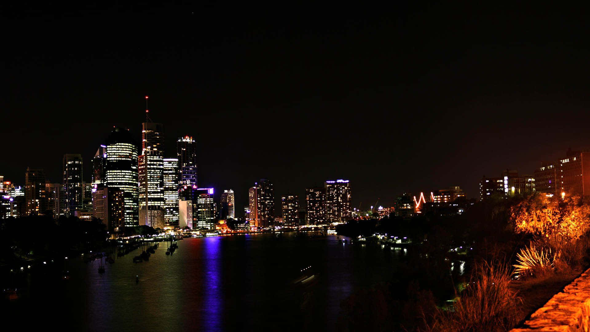 暗夜城市 桌布 壁紙 — 充滿活力的夜景城市景觀，摩天大樓在無星的天空下倒映在黑暗的河流上。