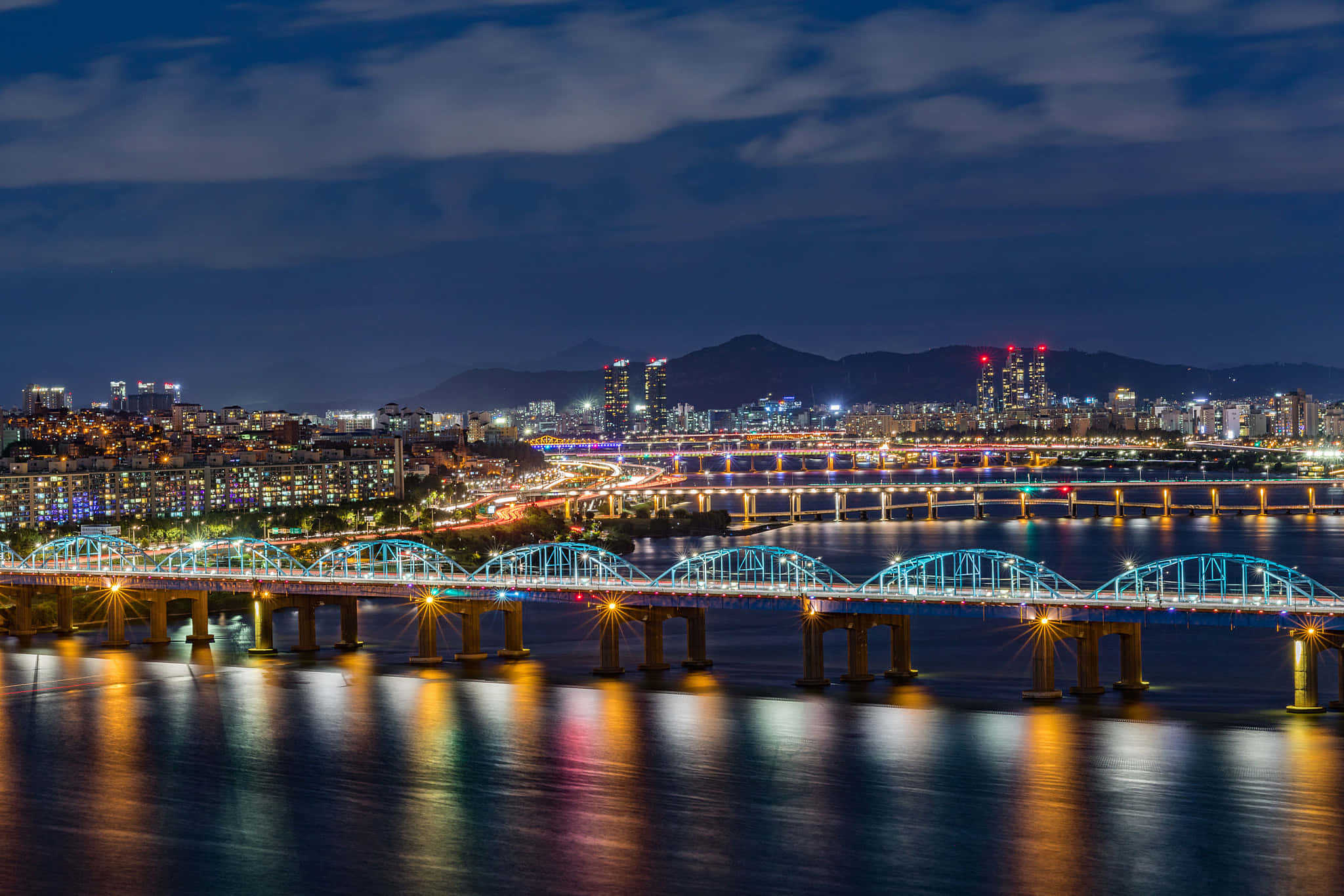 South Korea 桌布 壁紙 — 充滿活力的夜景城市景觀，特色是水面上燈火通明的橋樑，發光的燈光在水面反射，遠處的山脈在黑暗的天空下。