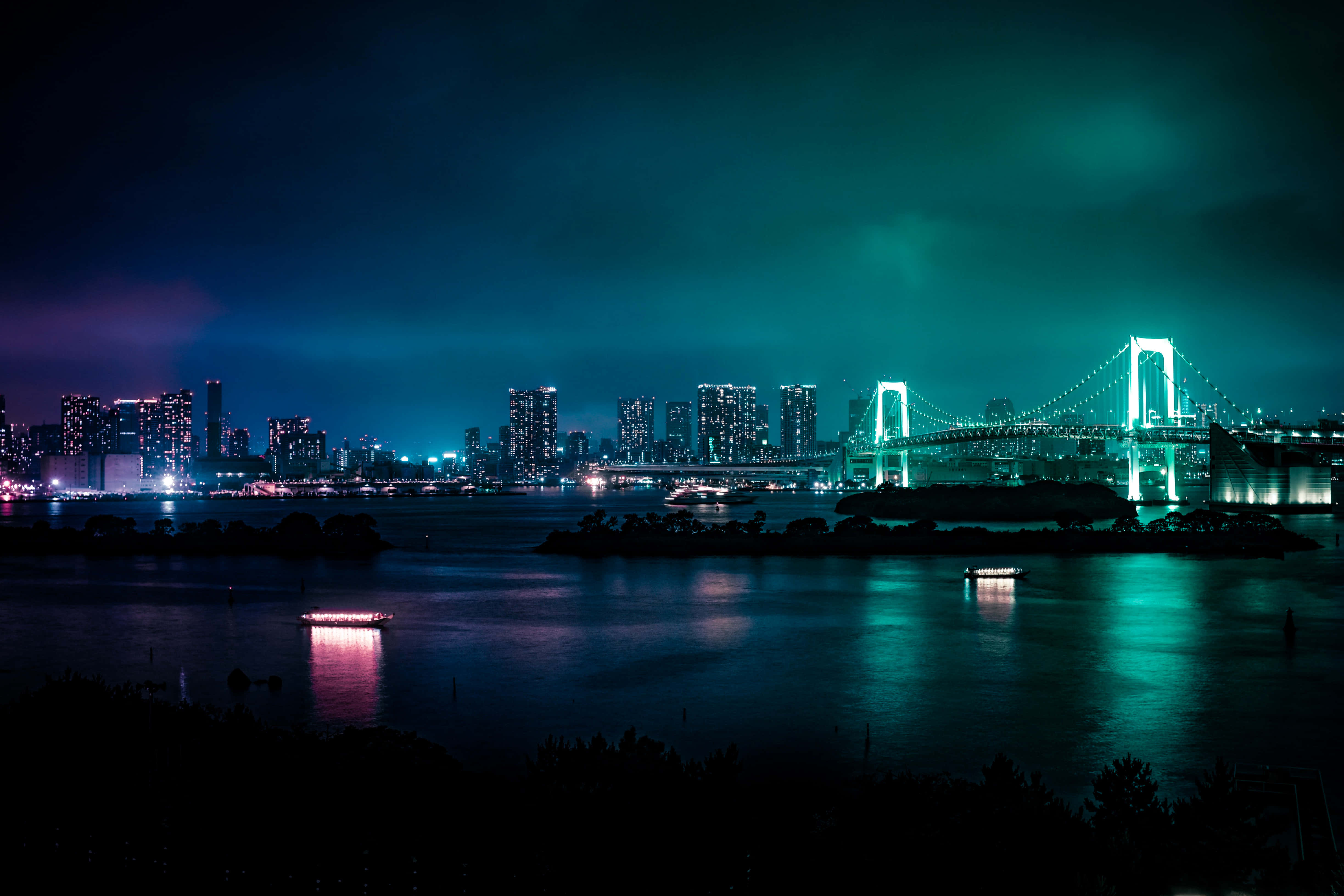 城市景觀 桌布 壁紙 — 充滿活力的夜景城市景觀，特色是發光的彩虹橋橫跨水面，有船隻和摩天大樓，在青綠色的天空下。