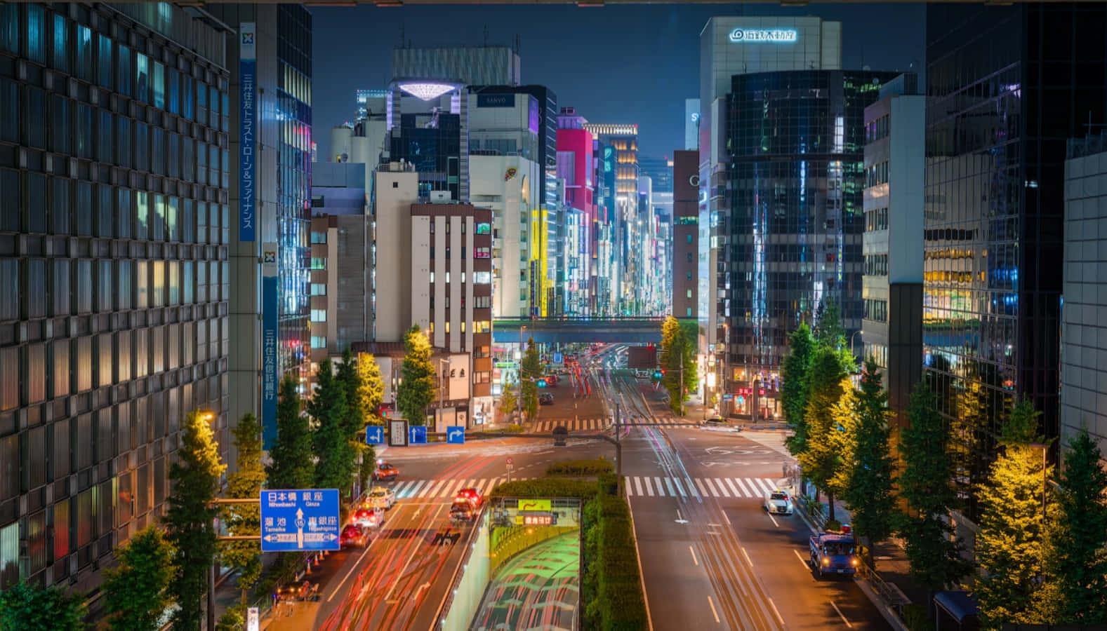 東京 桌布 壁紙 — 充滿活力的夜間城市景觀，閃爍的霓虹燈招牌和流動的車燈照亮了一條兩旁高樓大廈林立、繁華的都市街道。