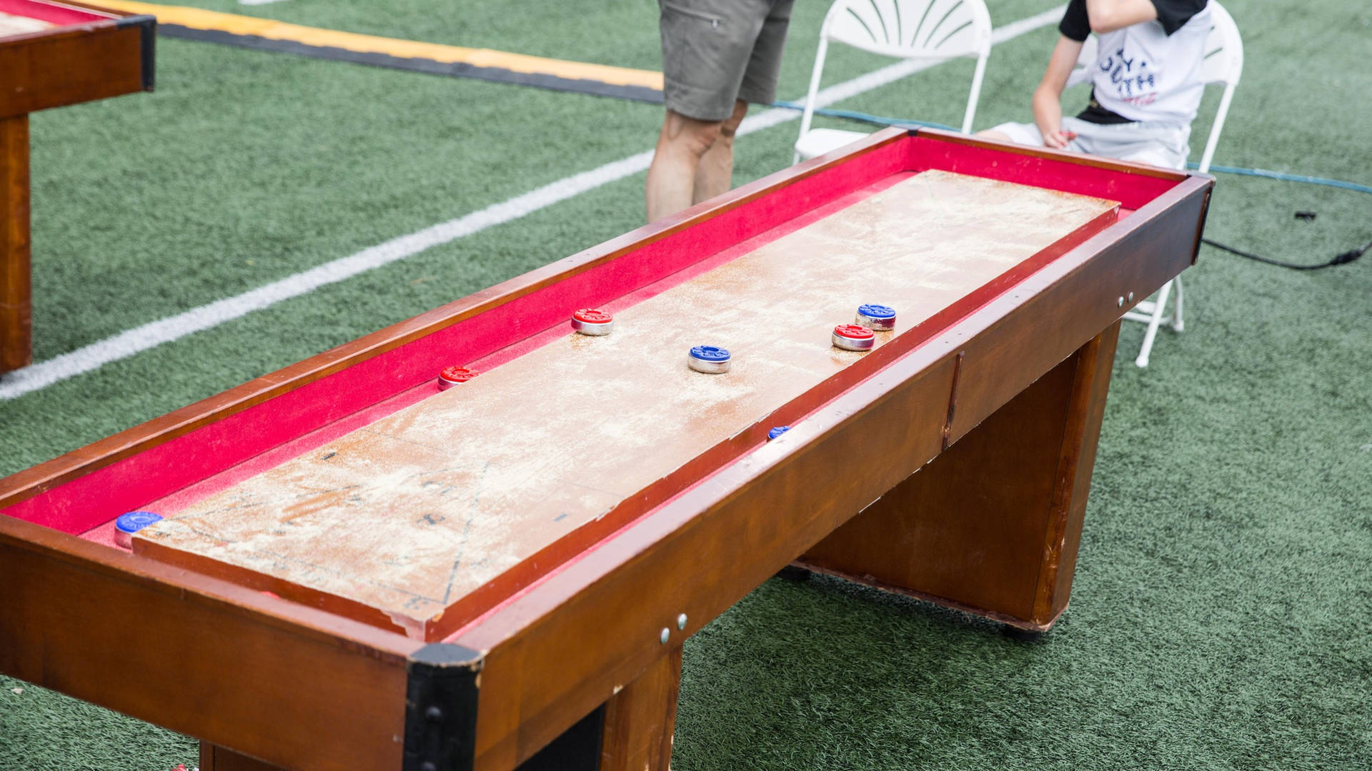 充滿活力的室內 Shuffleboard 遊戲進行中 壁紙 — 一張木製沙狐球桌，帶紅色邊框，散落著冰球，放置在綠色人造草皮上，玩家們聚集在附近。