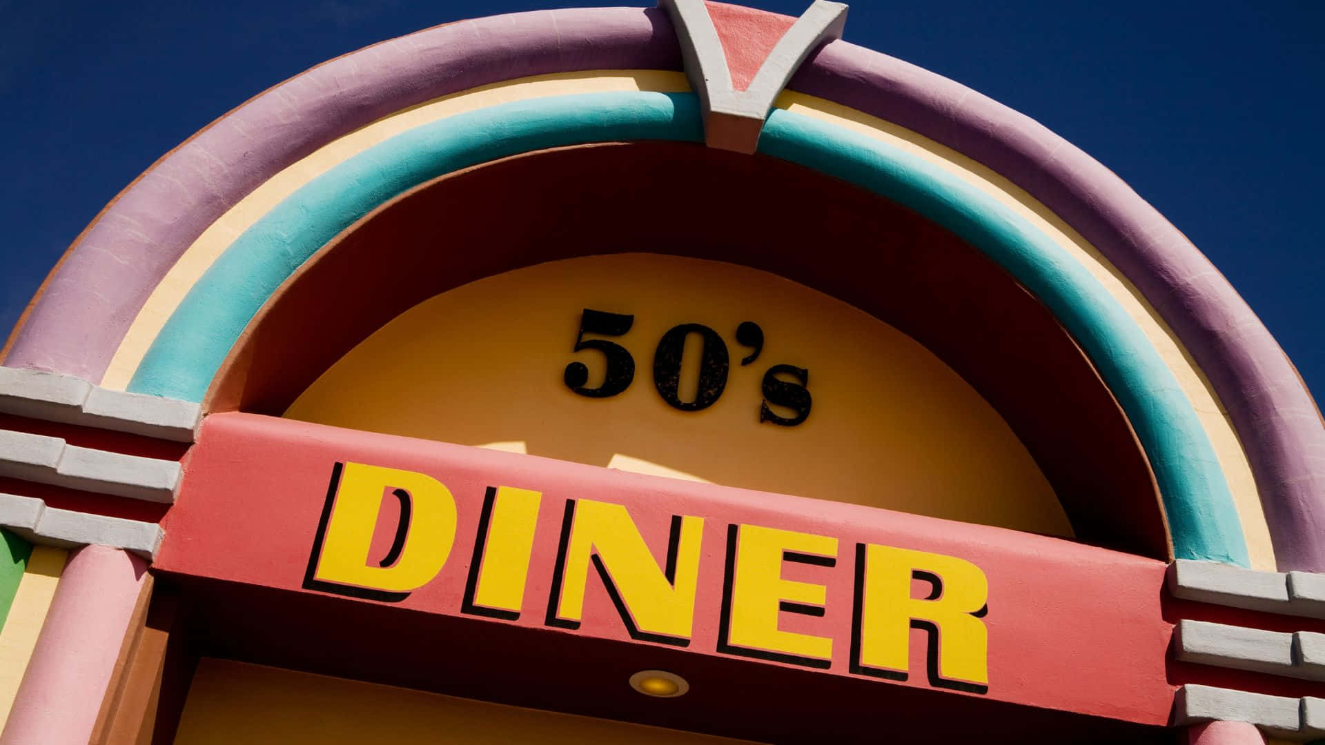 Diner 桌布 壁紙 — 充滿活力的復古餐廳招牌，帶有彩虹拱門和粗體黃色字體，襯托著清澈的藍天。