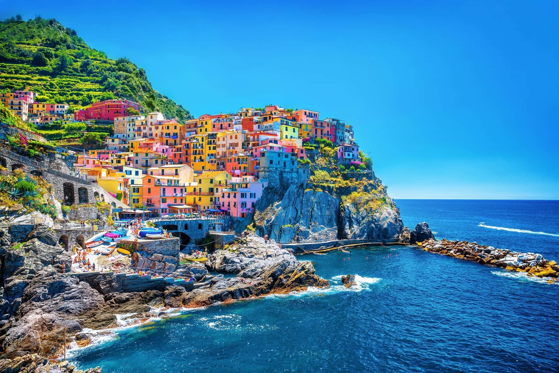 Italian Beach 桌布 壁紙 — 充滿活力的懸崖村莊，粉彩房屋沿著岩石地形層疊而下，旁邊是深藍色的大海，天空晴朗。