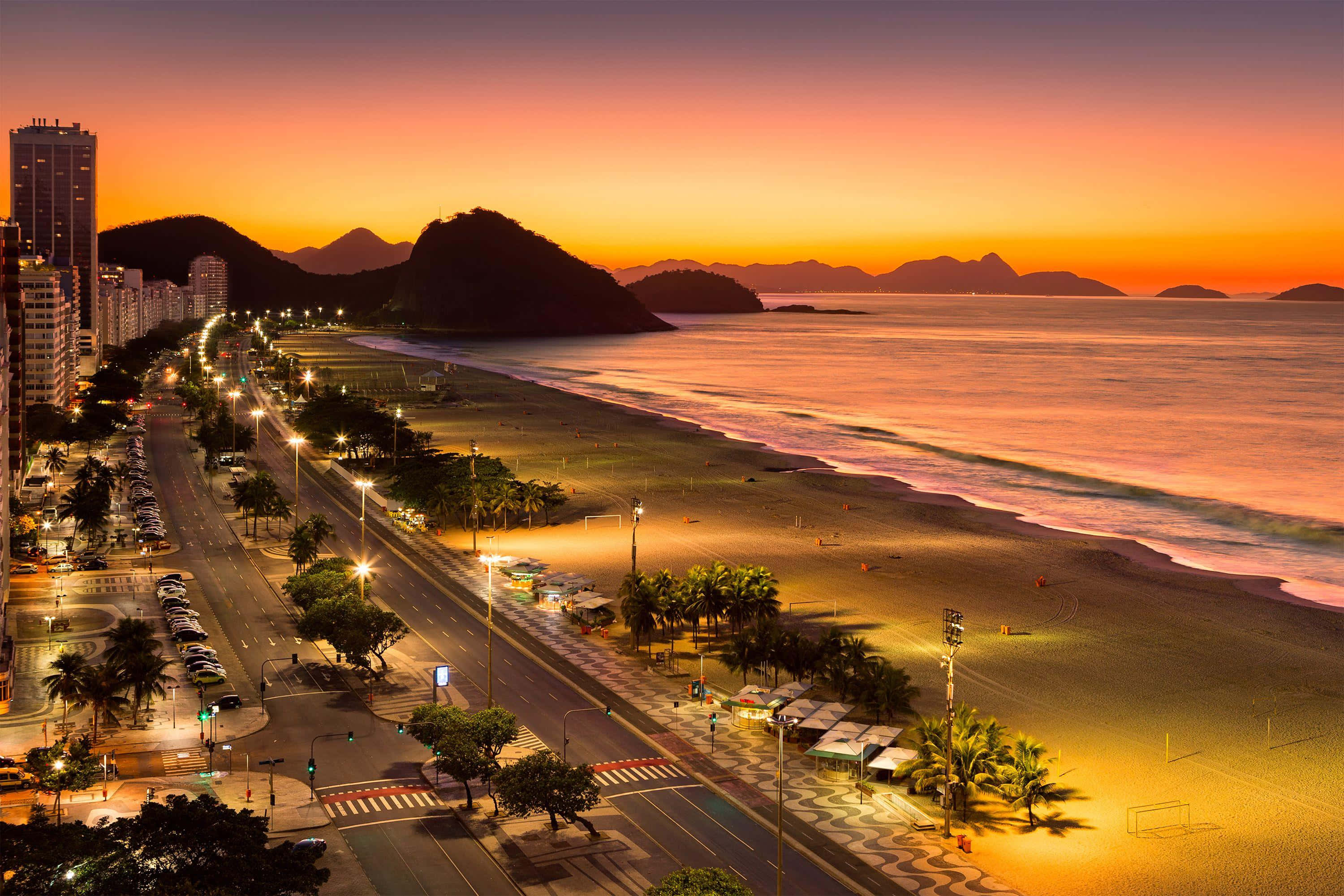 Brazil 桌布 壁紙 — 充滿活力的日落，照耀著繁華的海濱城市景觀，街道閃爍著光芒，棕櫚樹和遠山在橙、粉紅和紫色的天空中形成剪影。