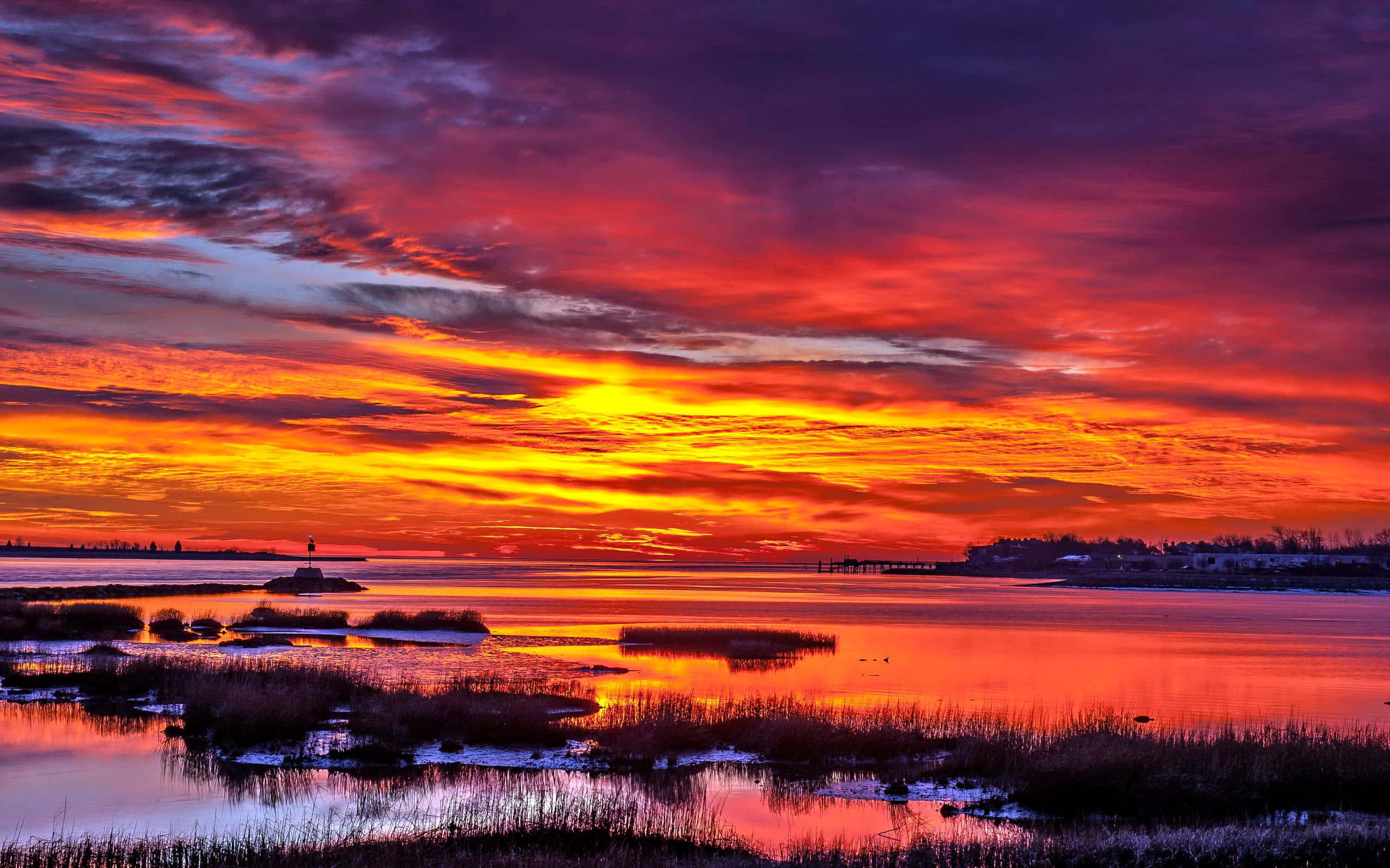 Sunset Landscape 桌布 壁紙 — 充滿活力的日落將天空染成火紅、橙色和紫色，在平靜的水面上生動地反射，遠處有燈塔和沼澤地。