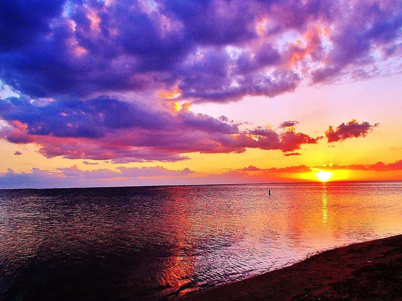 Beautiful Beach 桌布 壁紙 — 充滿活力的日落將天空染成紫色、橙色和黃色，倒映在沙灘附近平靜的海面上。