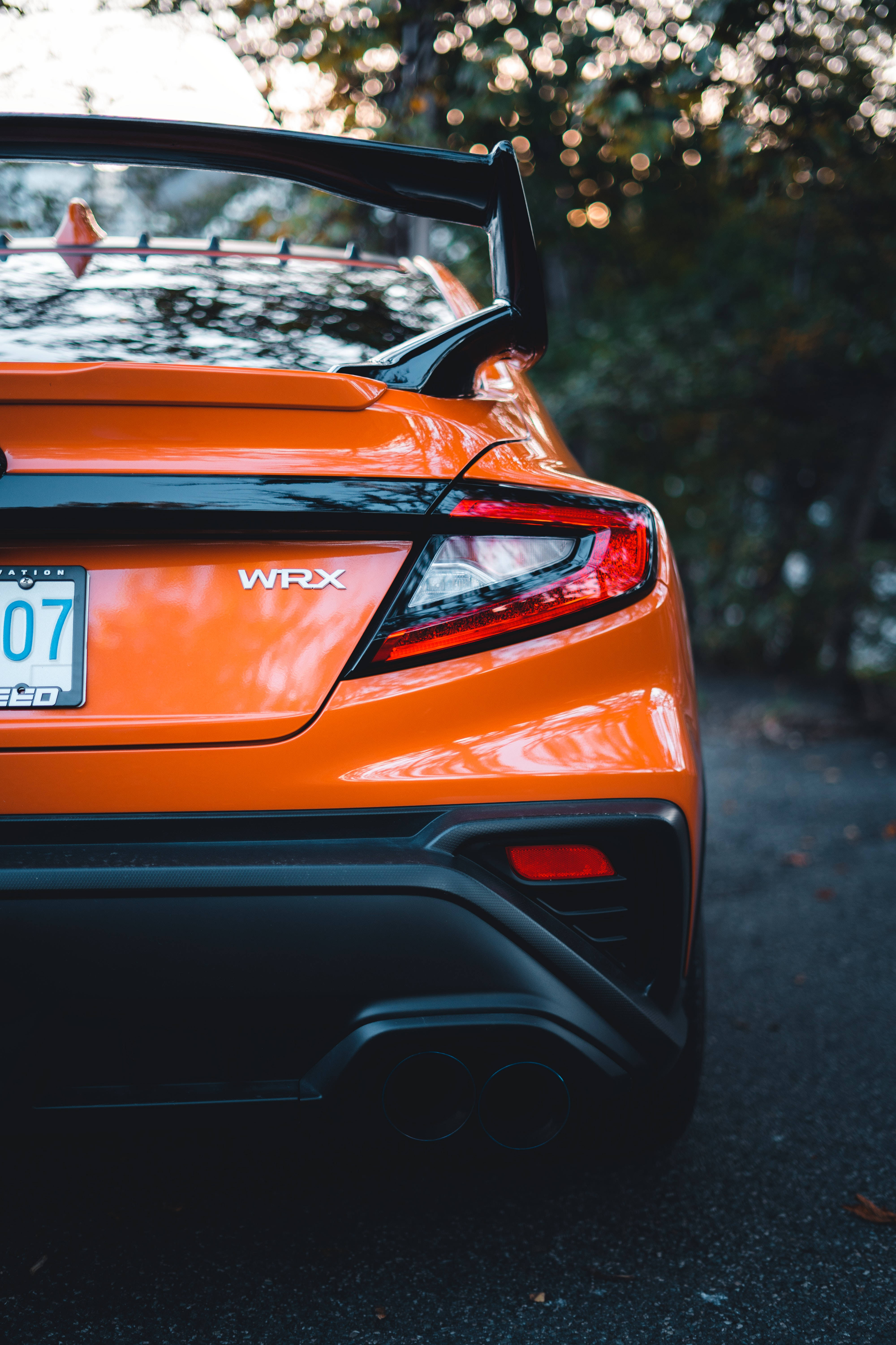 Sports Car 桌布 壁紙 — 充滿活力的橙色WRX跑車後視圖，配備黑色尾翼和雙出排氣管，停在斑駁陽光下的瀝青路面上。