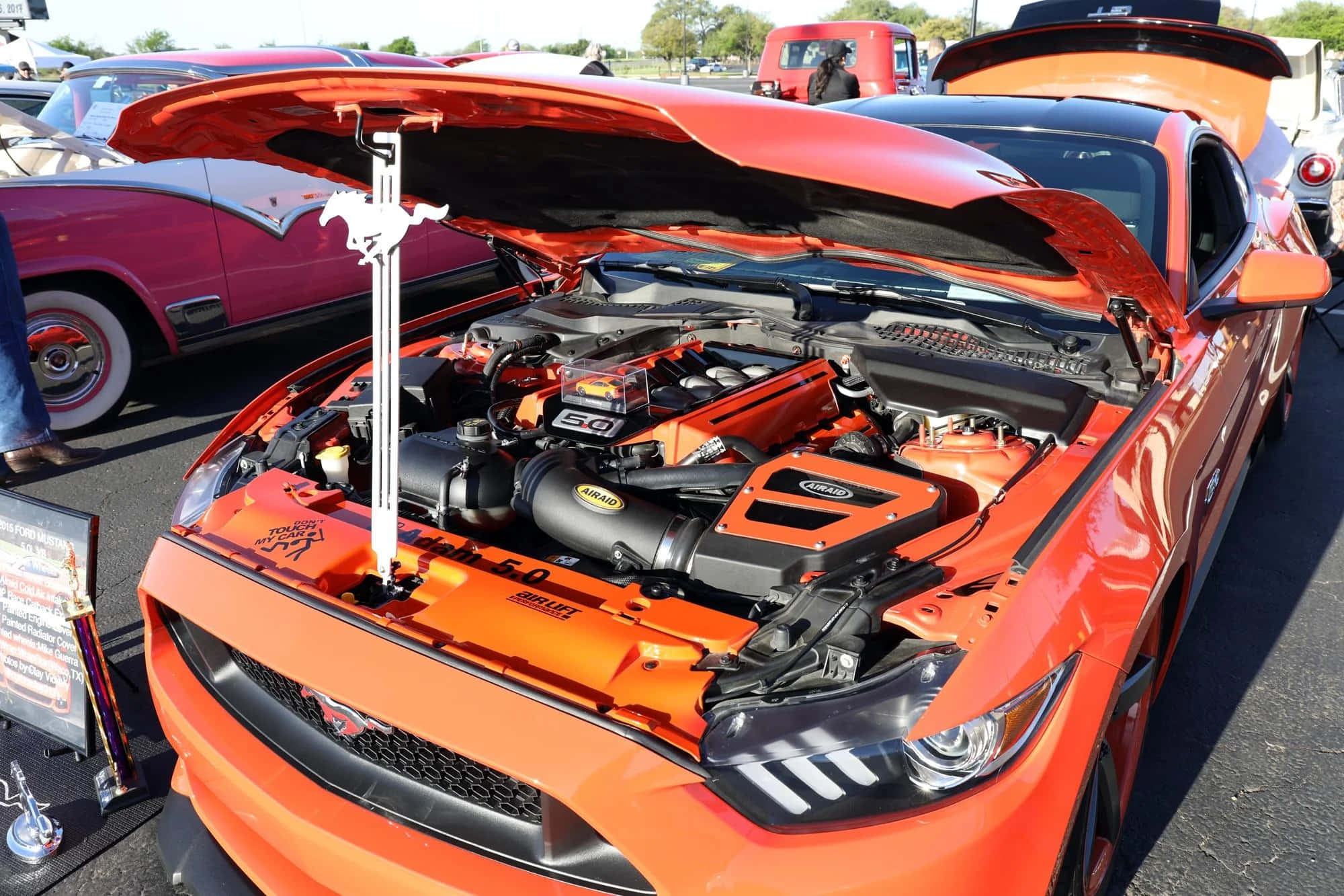 Car Hood 桌布 壁紙 — 充滿活力的橙色福特野馬，引擎蓋打開，在陽光明媚的車展上展示詳細的引擎。