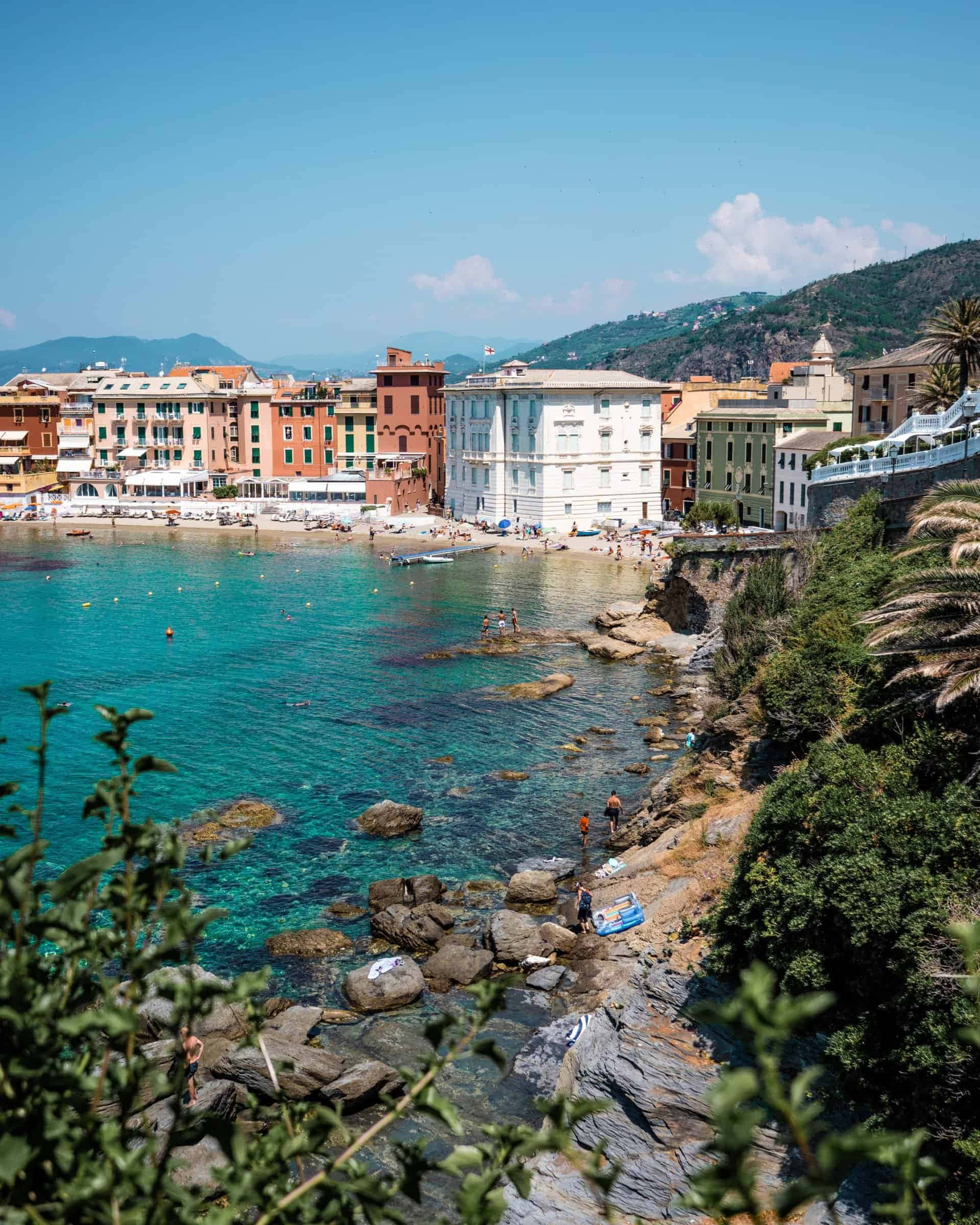 Italian Beach 桌布 壁紙 — 充滿活力的沿海小鎮，粉彩建築依偎在碧綠海水和岩石海岸旁，天空清澈湛藍。