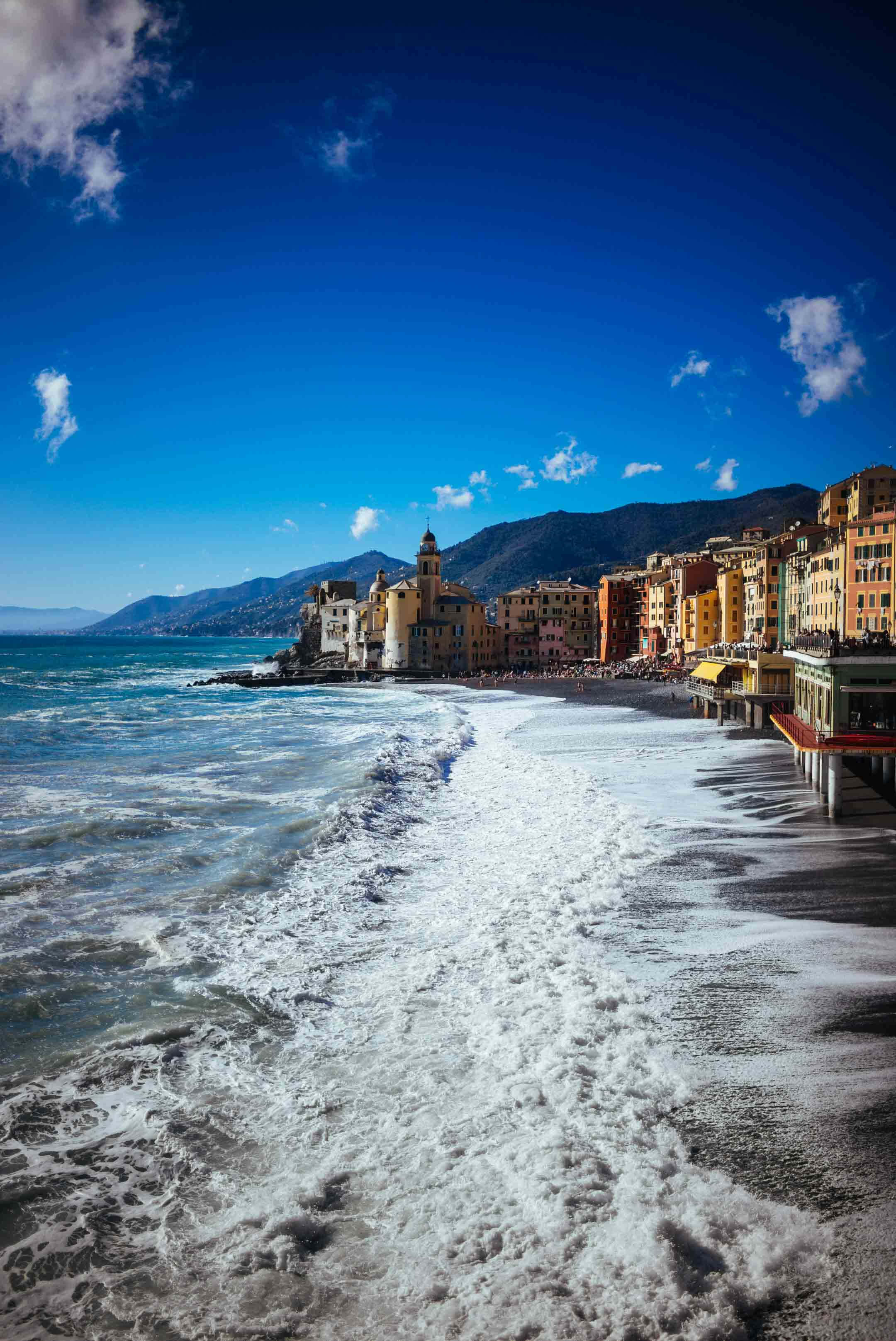 Italian Beach 桌布 壁紙 — 充滿活力的沿海小鎮，色彩繽紛的建築物擁抱著沙灘，海浪在清澈的藍天下拍打。