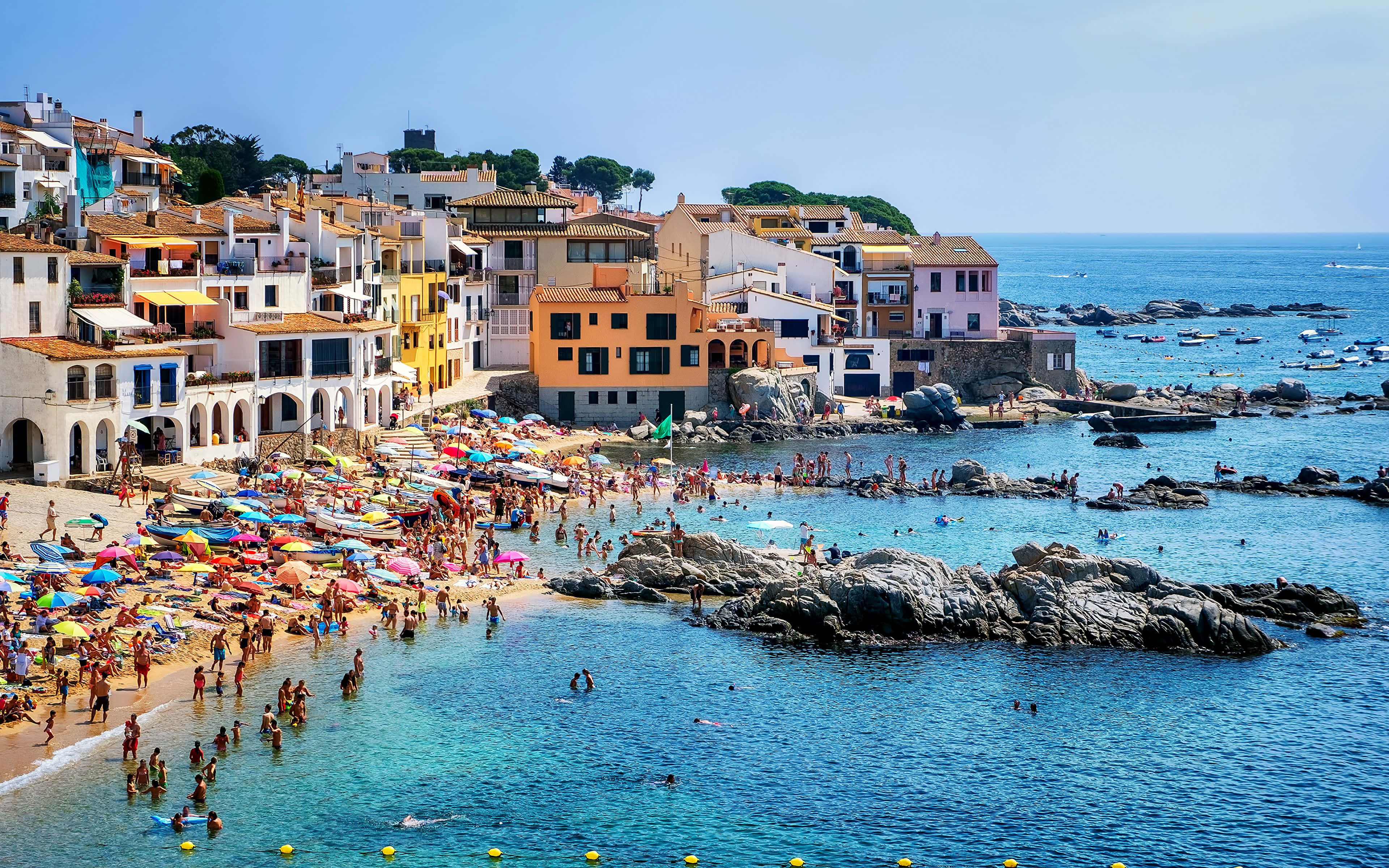 Spanish Beach 桌布 壁紙 — 充滿活力的沿海小鎮，色彩繽紛的建築物坐落在懸崖上，俯瞰著擠滿曬太陽和游泳人士的熱鬧海灘，海水呈碧綠色。