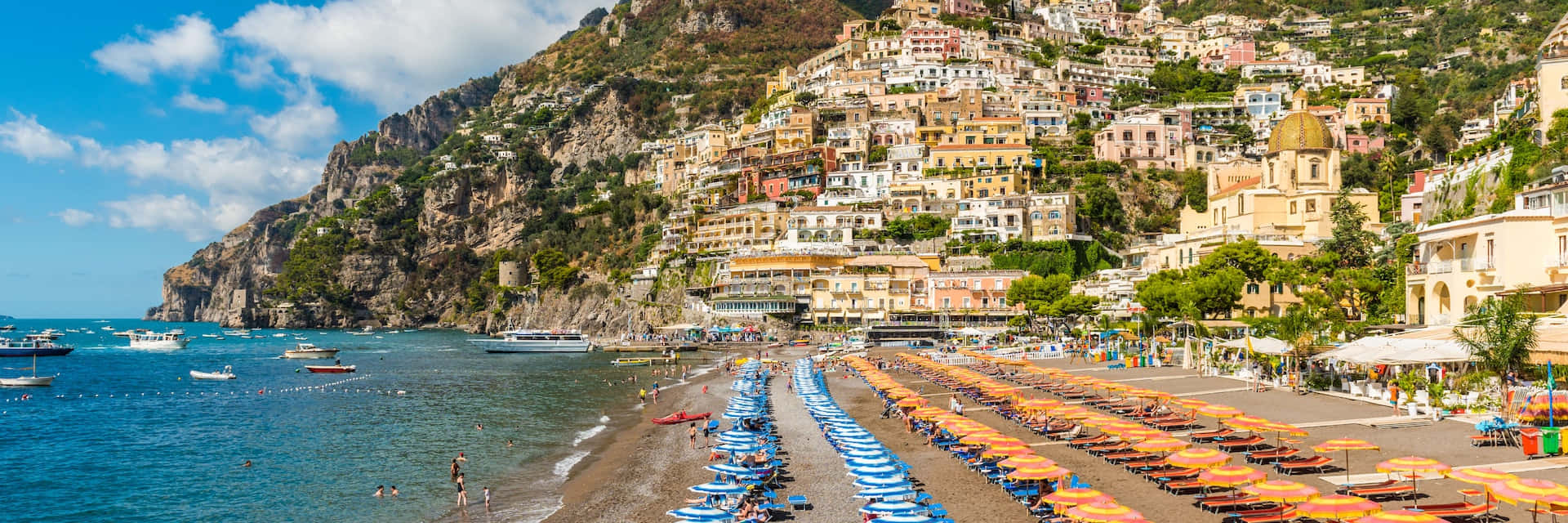 Italian Island 桌布 壁紙 — 充滿活力的沿海小鎮沿著陽光普照的海灘旁的懸崖傾瀉而下，海灘上排列著藍色和橙色的遮陽傘。