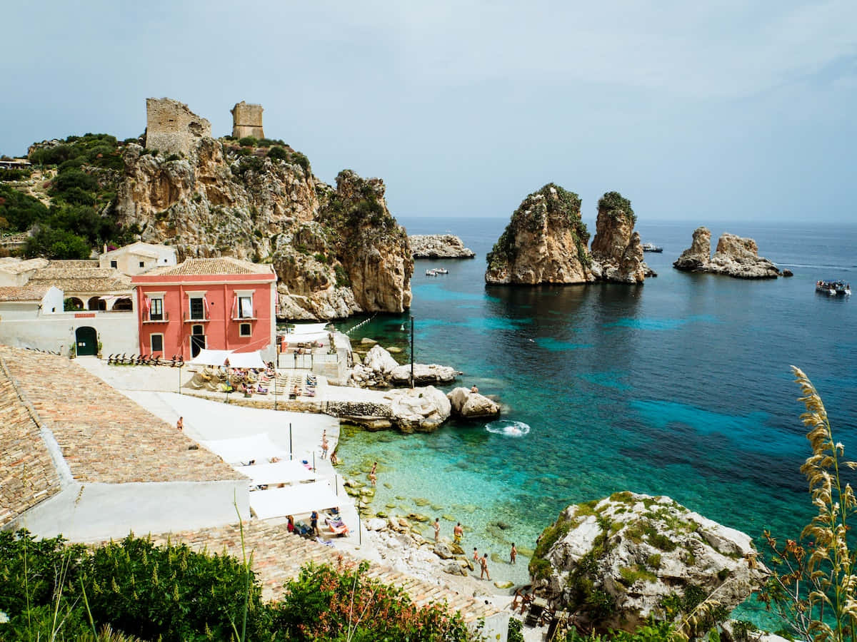 Italian Island 桌布 壁紙 — 充滿活力的海岸景色，有碧綠的海水、岩石峭壁，以及靠近沙灘的紅色建築，人們在享受陽光。