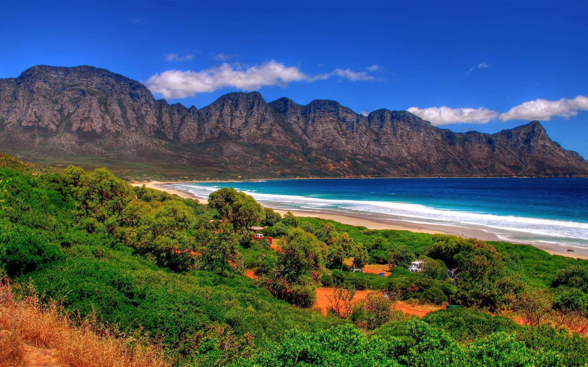 South Africa 桌布 壁紙 — 充滿活力的海岸景色，綠松石色的海浪拍打著沙灘，四周環繞著鬱鬱蔥蔥的綠色山丘和巍峨崎嶇的山脈，天空湛藍清澈。