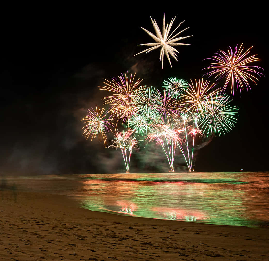 新年動畫 桌布 壁紙 — 充滿活力的煙火在夜間海灘上空爆炸，在水面和沙灘上投下色彩繽紛的倒影。