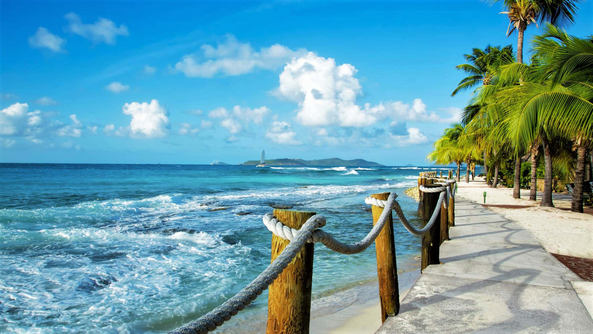 Caribbean 桌布 壁紙 — 充滿活力的熱帶海灘場景，青綠色的海浪拍打著木製繩索欄杆，湛藍的天空點綴著蓬鬆的白雲。
