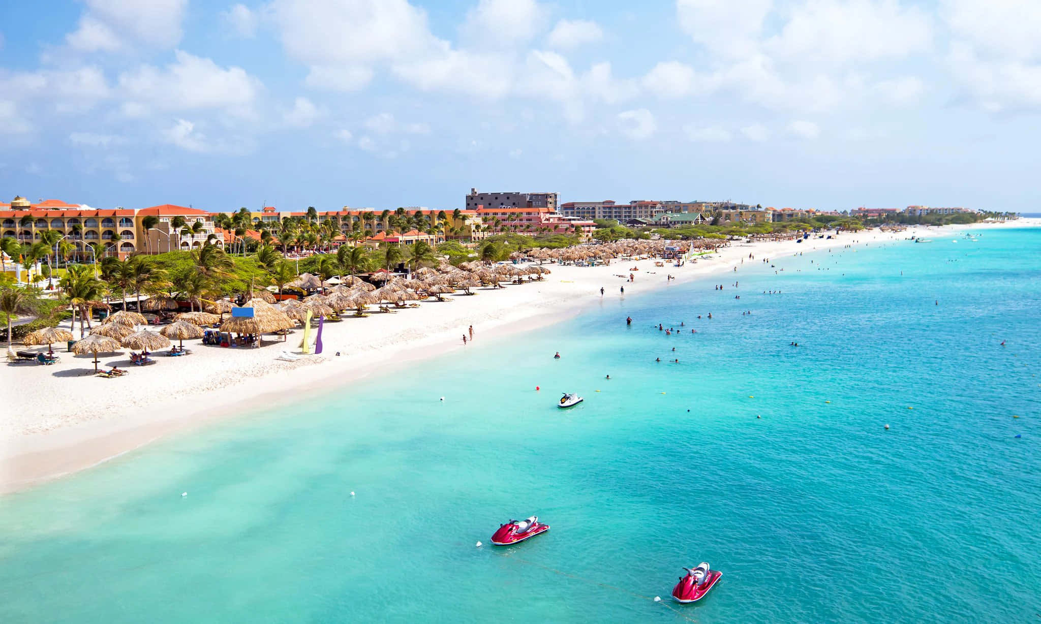 Aruba 桌布 壁紙 — 充滿活力的熱帶海灘鳥瞰圖，碧綠海水、水上電單車和度假村建築在明亮的藍天下。