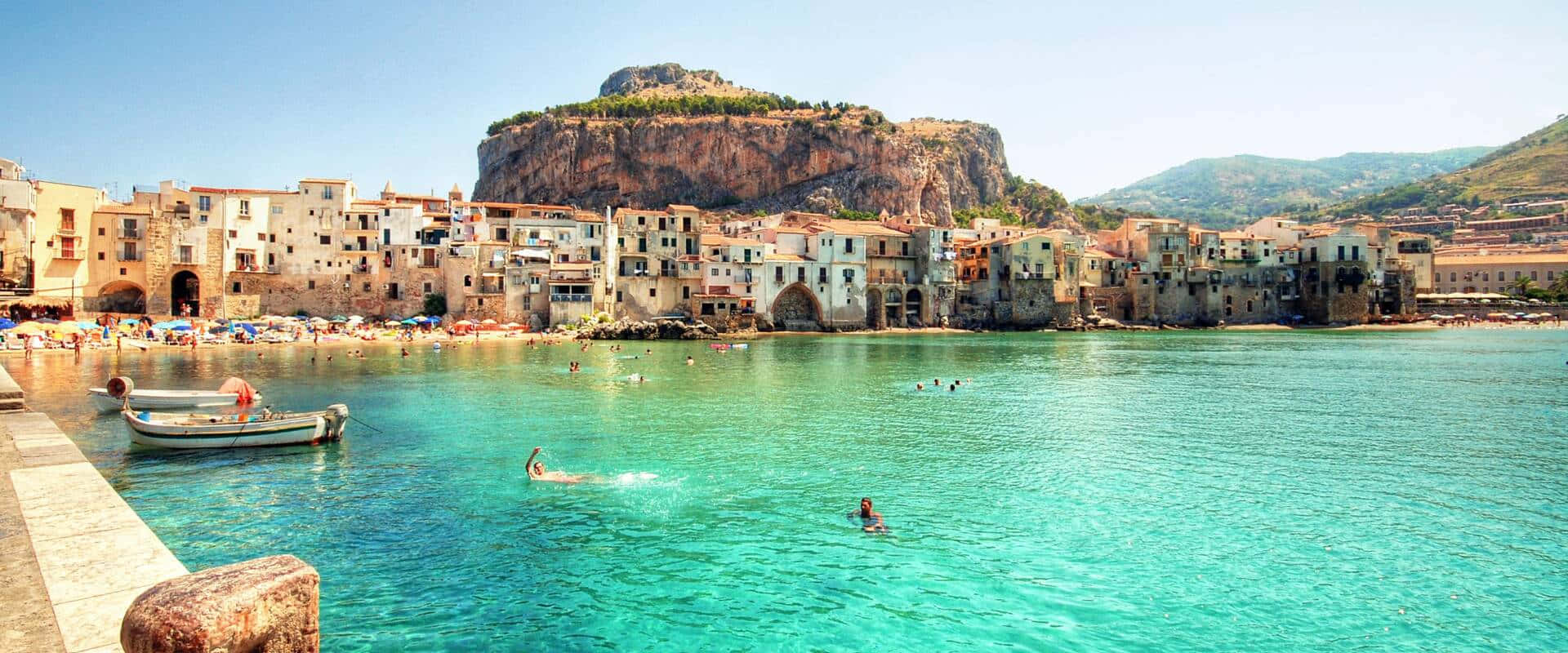 Italian Island 桌布 壁紙 — 充滿活力的碧綠海水拍打著陽光明媚、依附在岩石懸崖上的沿海小鎮，人們在游泳，船隻停泊在碼頭。