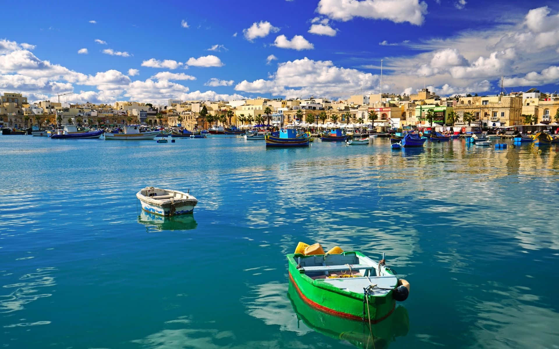 Malta 桌布 壁紙 — 充滿活力的碧綠海水映照著明亮的藍天，色彩繽紛的漁船漂浮在陽光普照的沿海小鎮附近，那裏有陶土屋頂的建築。