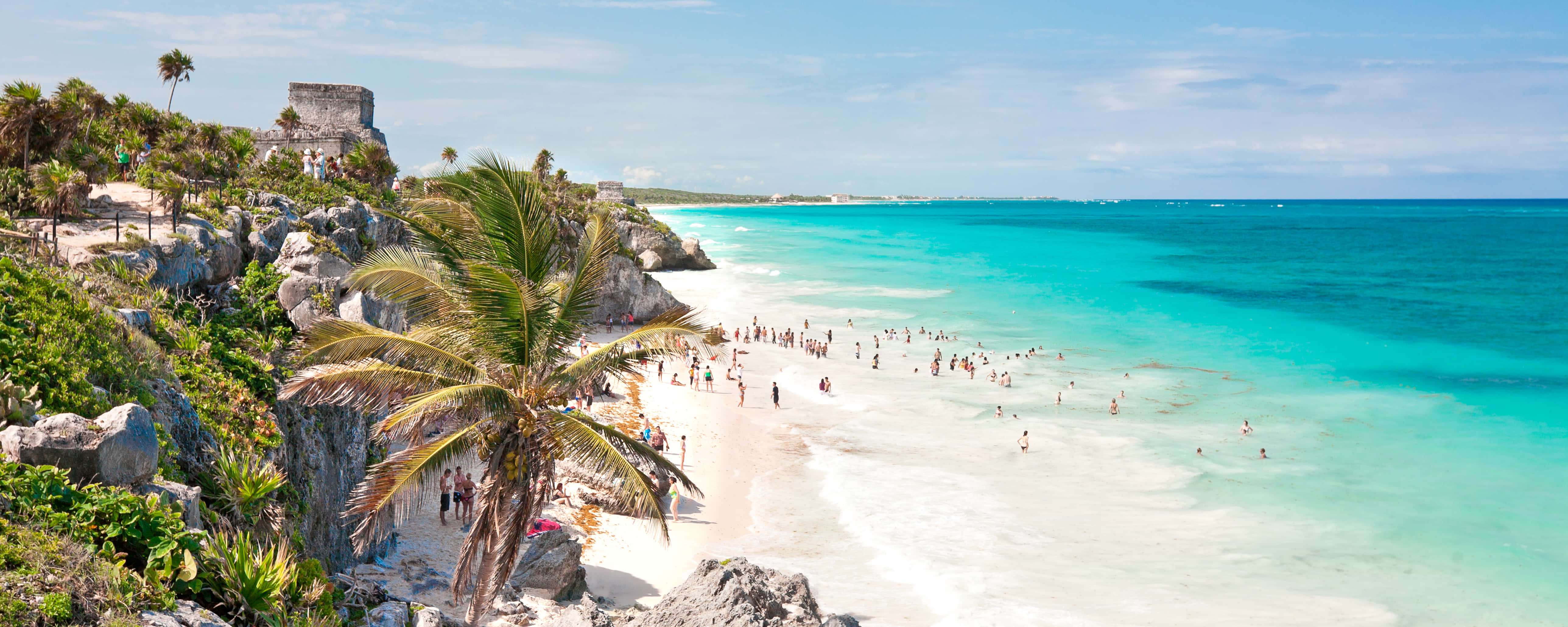 Mexican Beach 桌布 壁紙 — 充滿活力的碧綠海洋與白色沙灘相遇，沙灘上擠滿了享受日光浴的人們，頭頂是明亮的藍天，岩石峭壁上矗立著古老的遺跡。