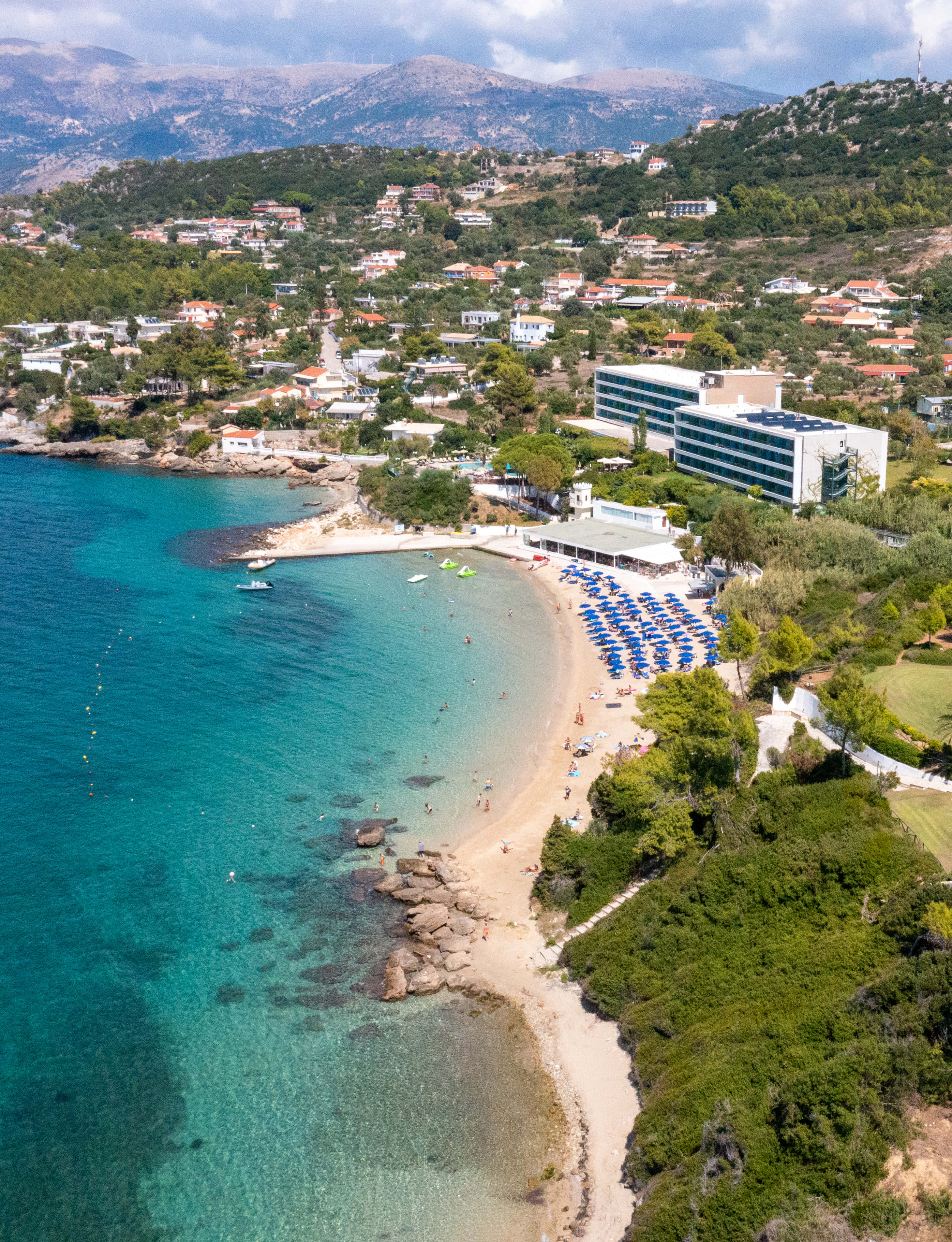 Mediterranean Beach 桌布 壁紙 — 充滿活力的碧綠海灣鳥瞰圖，沙灘上排列著藍色遮陽傘，周圍環繞著綠色山丘和度假村建築。