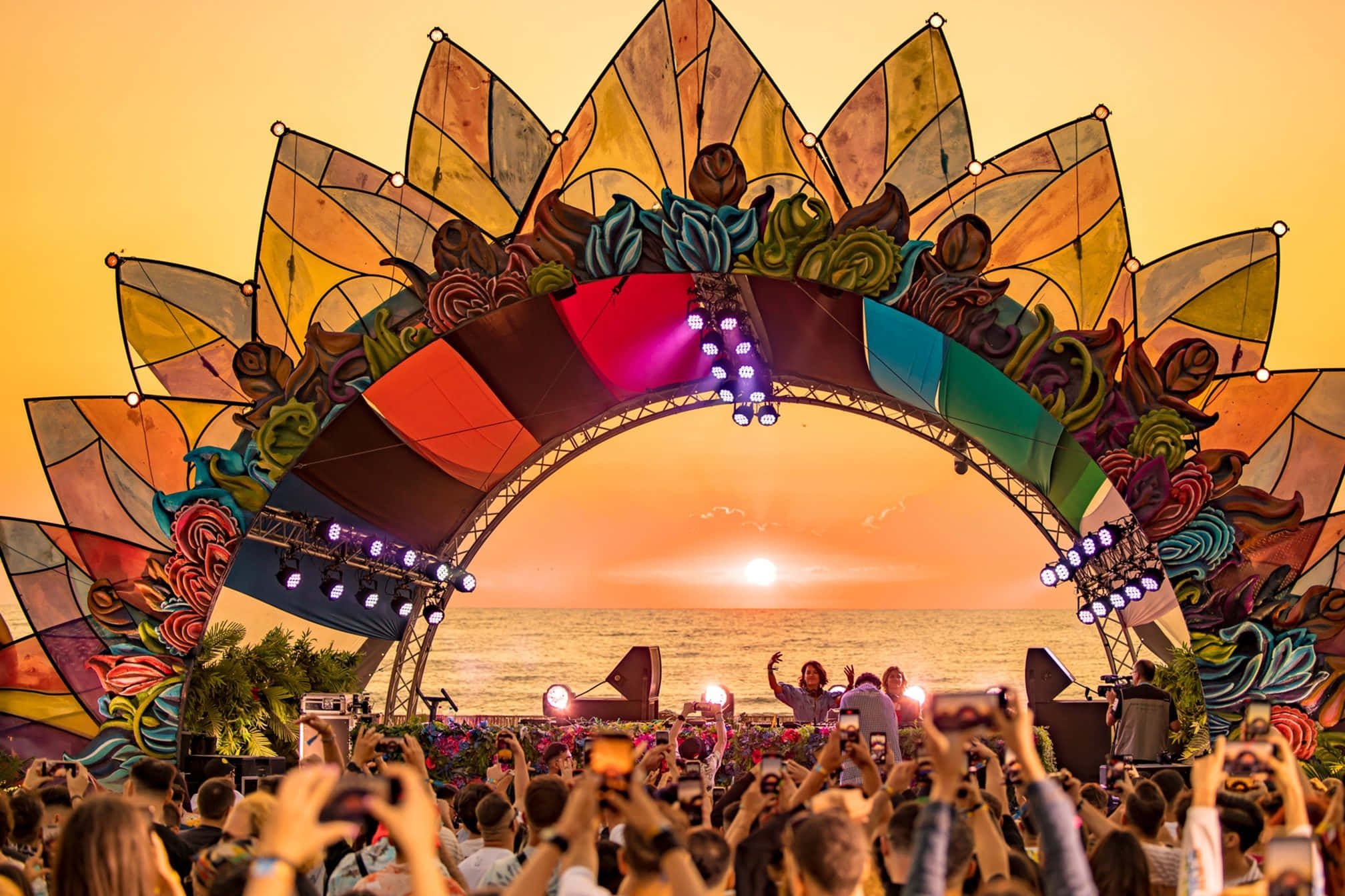 Beach Party 桌布 壁紙 — 充滿活力的節日人群透過色彩繽紛的彩繪玻璃拱門捕捉日落，拱門俯瞰著海洋。