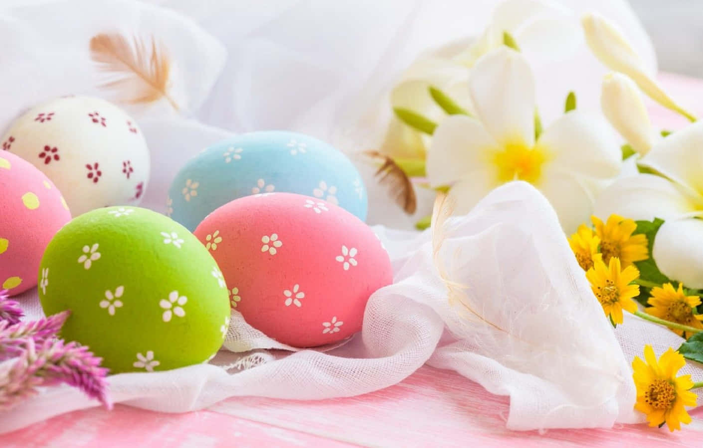 Pastel Easter 桌布 壁紙 — 充滿活力的粉彩復活蛋帶有花卉圖案，放在白色布料上，旁邊有黃色花朵和柔軟羽毛。