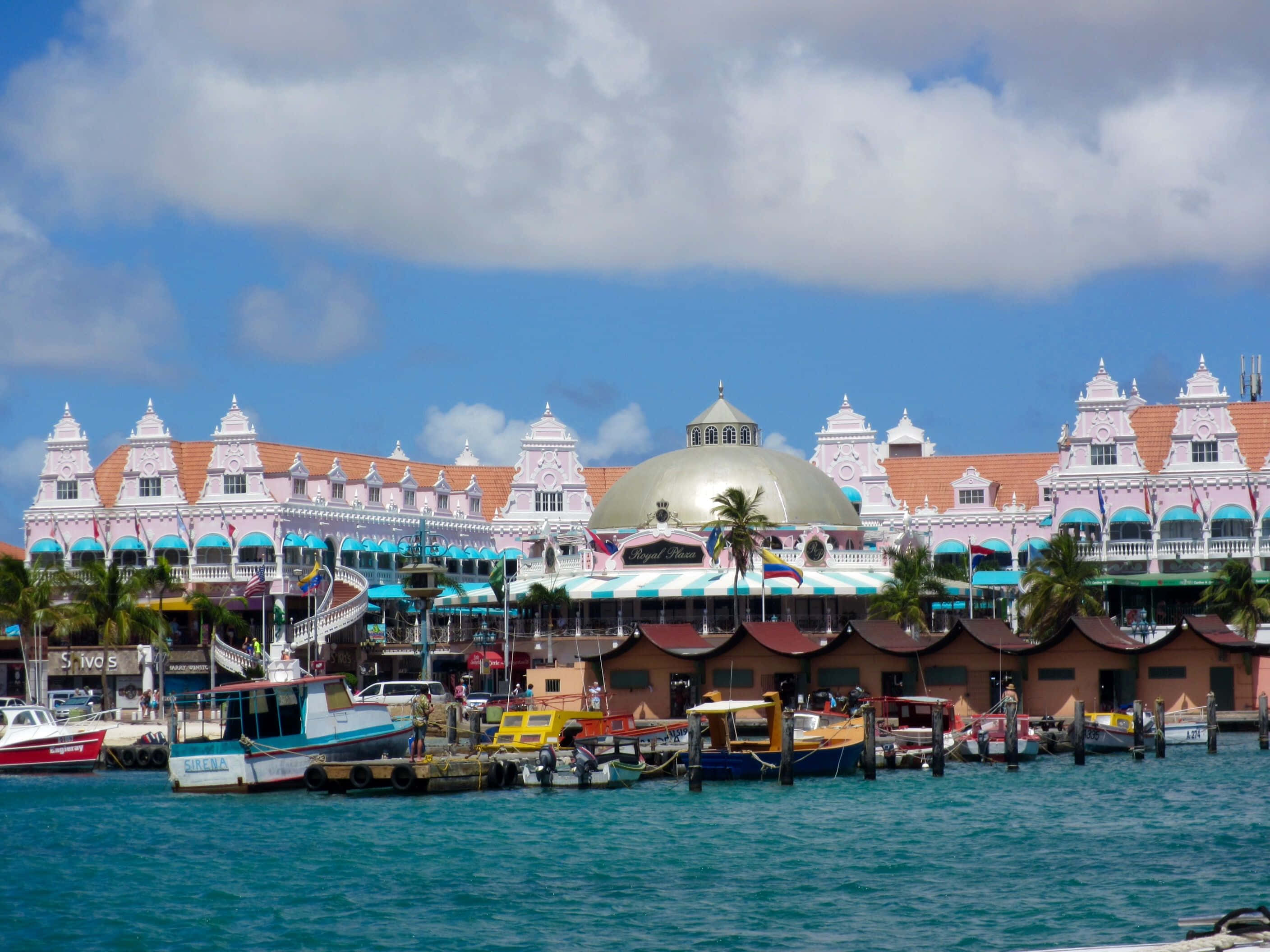 Aruba 桌布 壁紙 — 充滿活力的粉紅色度假村建築，配有藍綠色遮陽篷，排列在藍綠色的港口，港口裡停滿了色彩繽紛的船隻，上方是明亮的藍天和蓬鬆的雲朵。