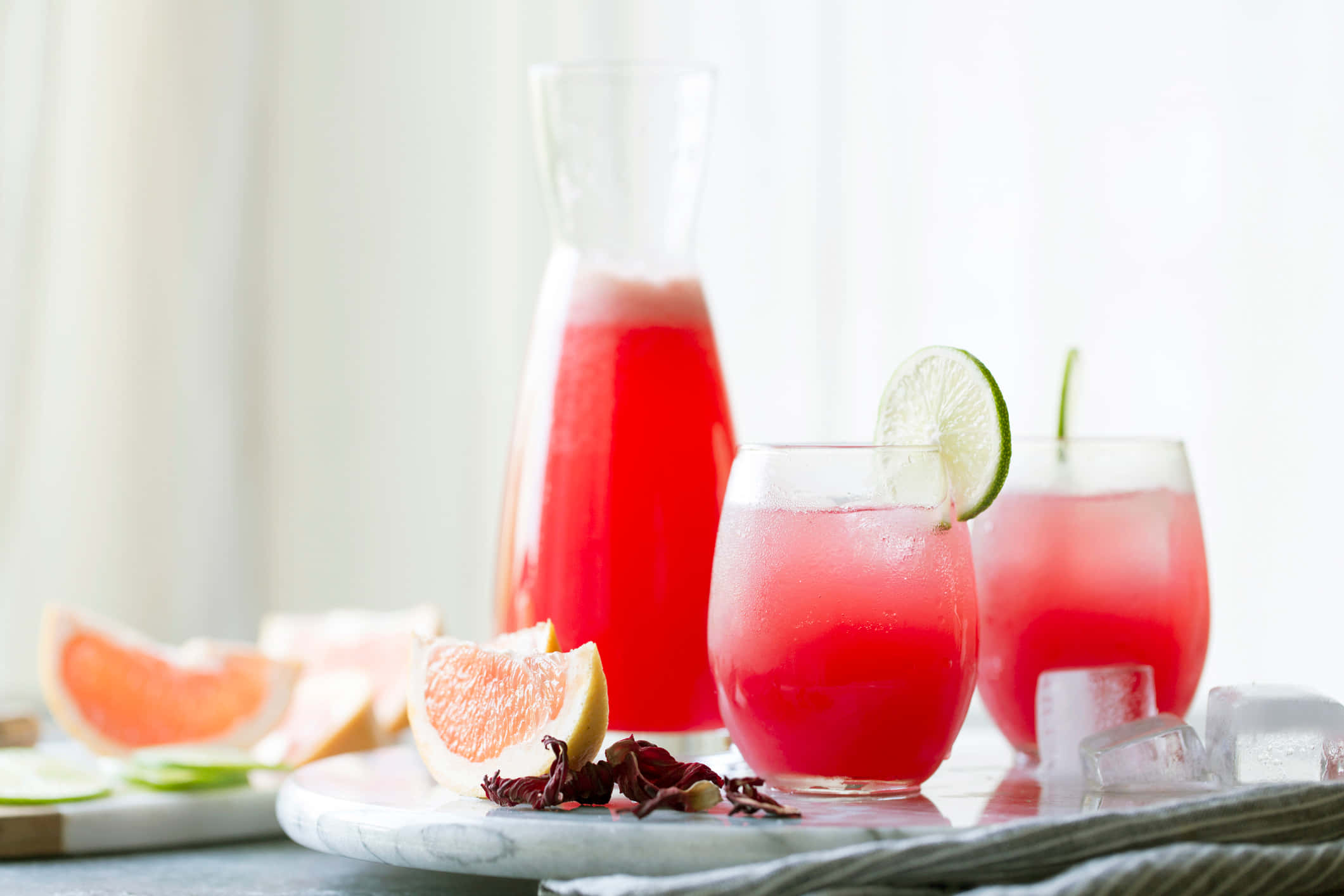 Beautiful Drink 桌布 壁紙 — 充滿活力的粉紅色木槿花飲料，配上青檸裝飾和冰塊，盛在玻璃杯中，旁邊有切片西柚和一個玻璃壺。