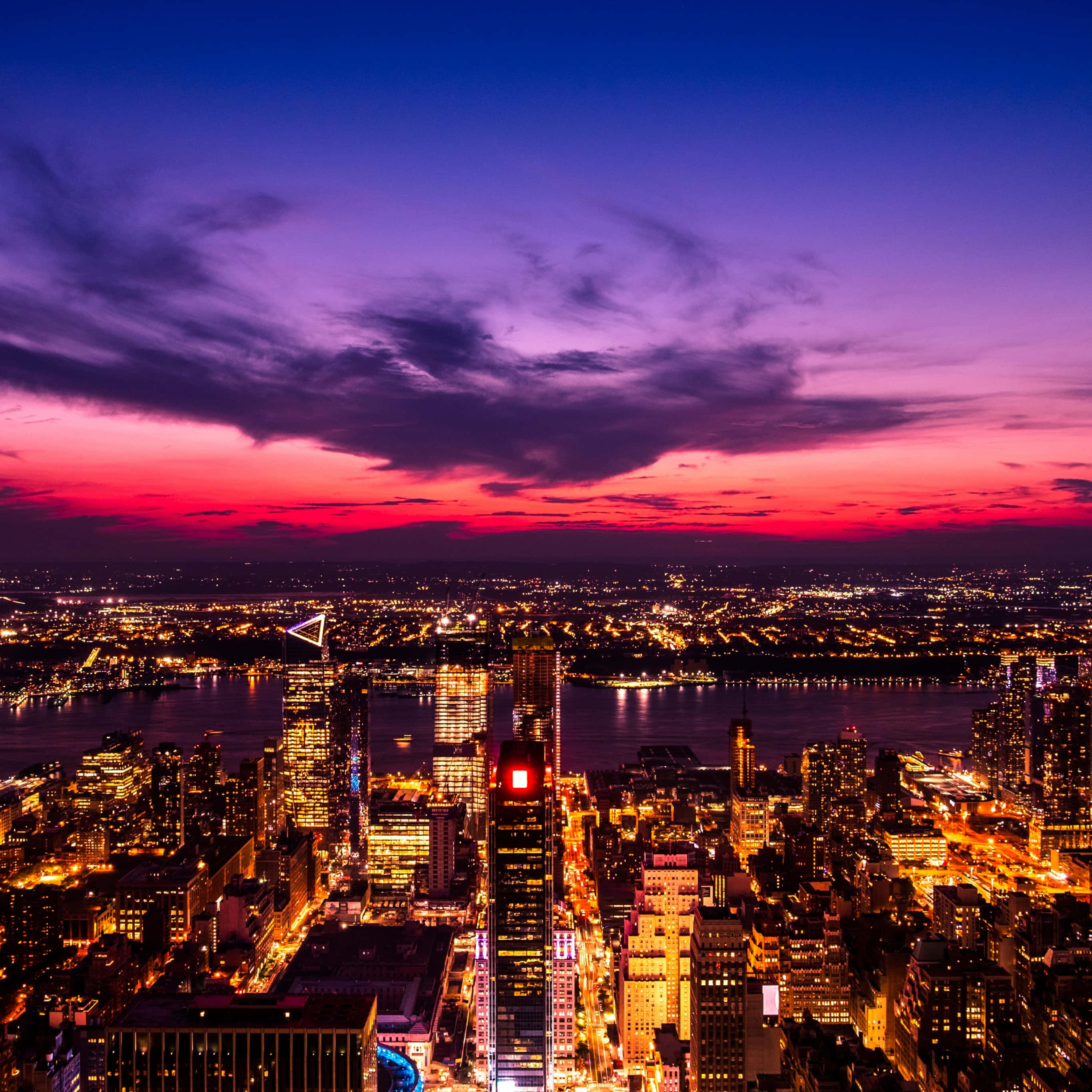 City Lights 桌布 壁紙 — 充滿活力的紫色和粉紅色日落照亮了廣闊的城市景觀，閃爍的摩天大樓和下方的河流。