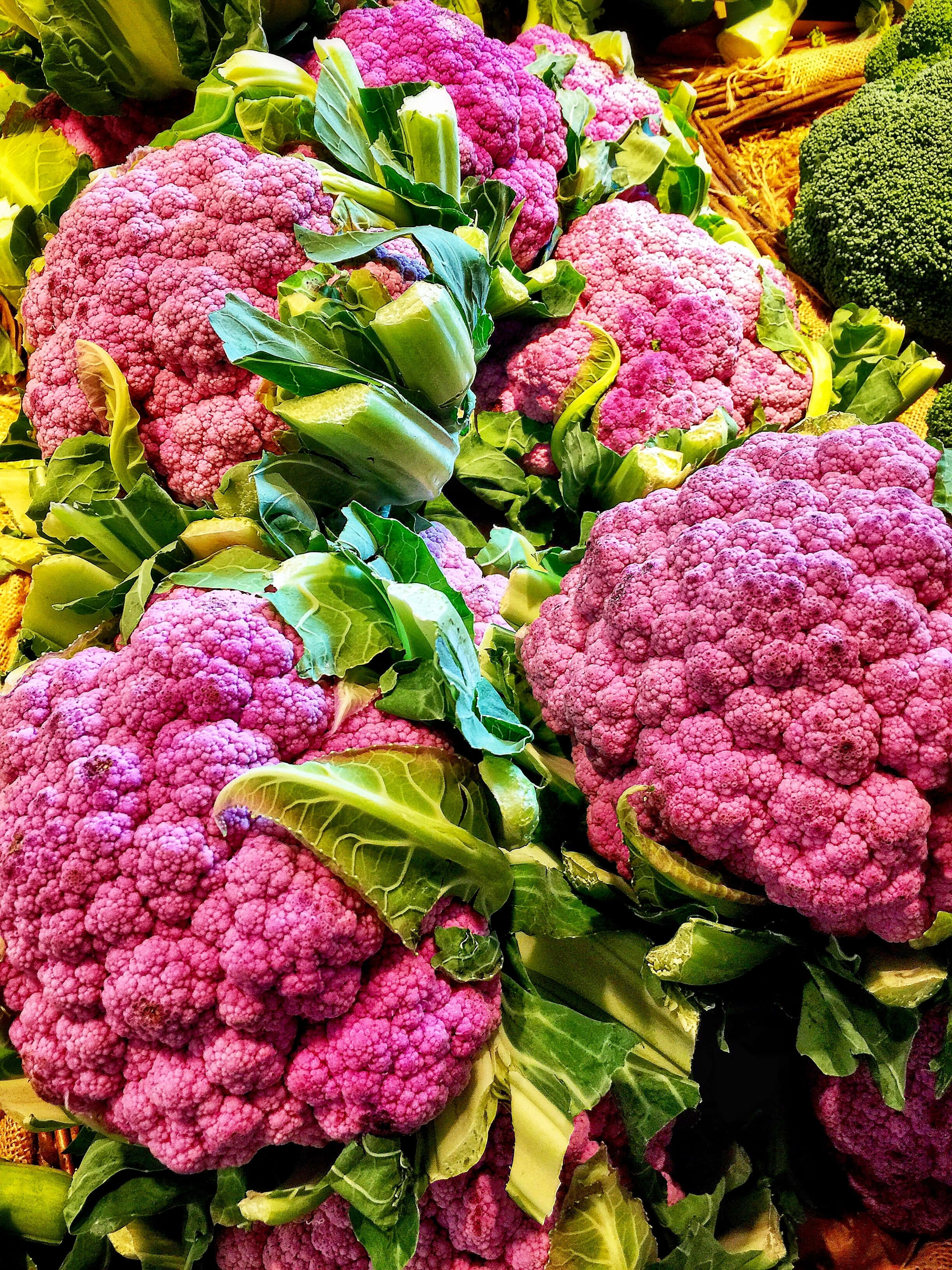 Purple Cauliflower 桌布 壁紙 — 充滿活力的紫色椰菜花頭，搭配茂盛的綠色葉子，堆疊在色彩繽紛的市場展示中。