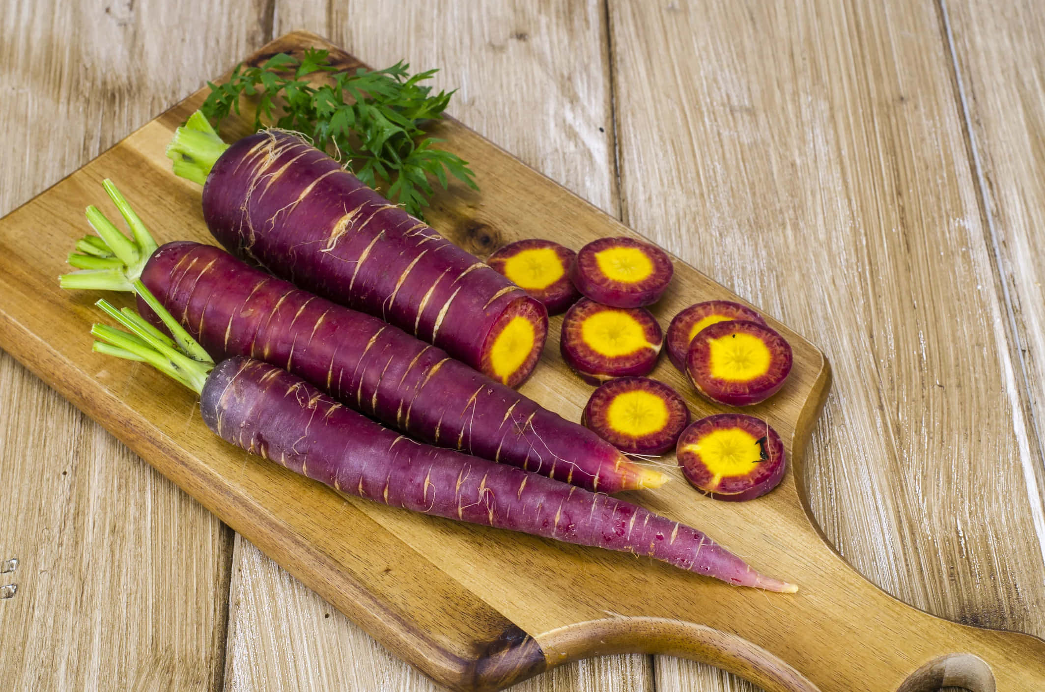 Purple Carrots 桌布 壁紙 — 充滿活力的紫色胡蘿蔔帶有明亮的黃色中心，放在質樸的木製砧板上，旁邊是新鮮的歐芹。