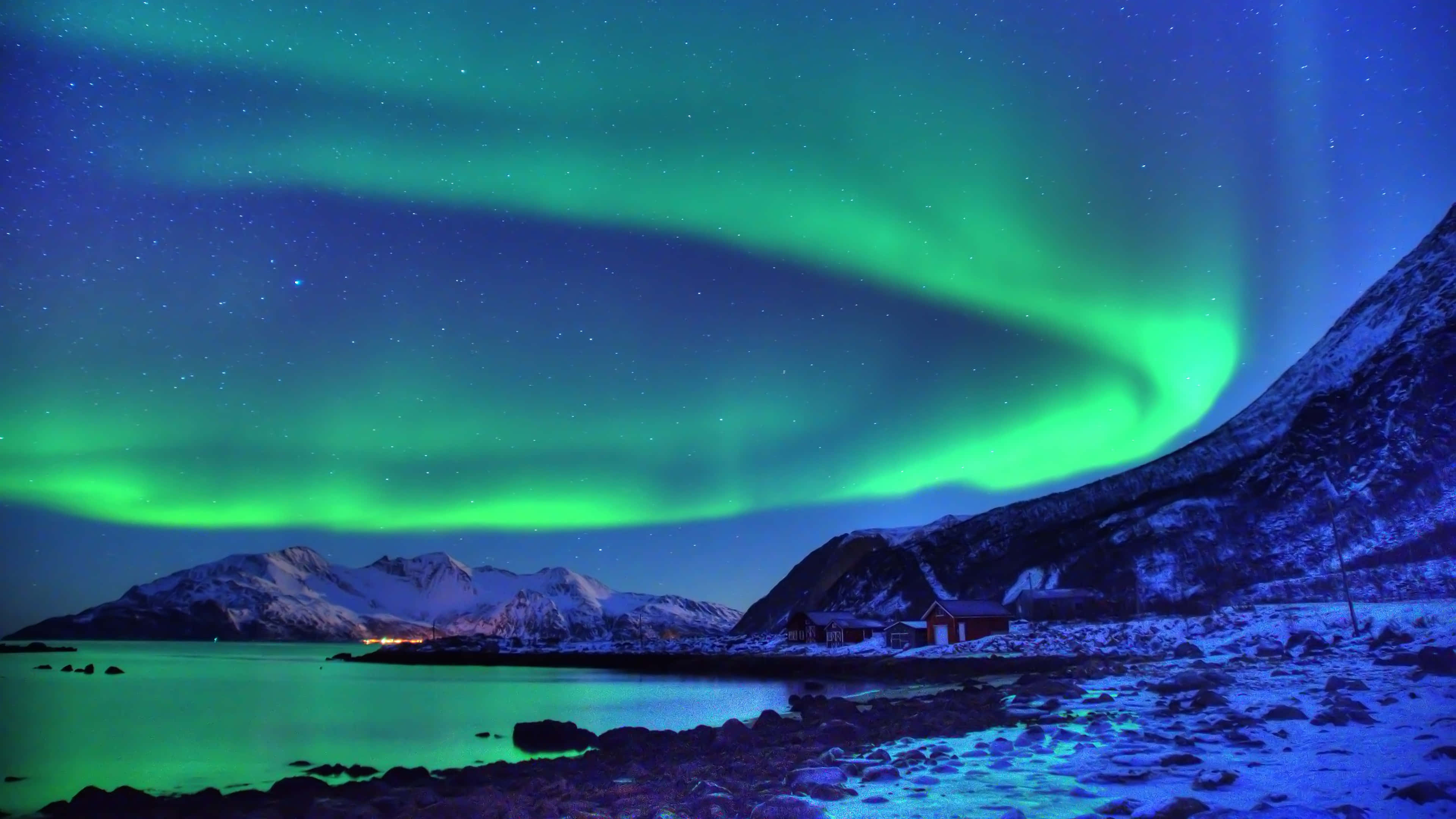 aurora borealis 桌布 壁紙 — 充滿活力的綠色北極光在繁星點點的夜空中舞動，映照著雪山和寧靜的峽灣，峽灣旁有一座紅色小屋。