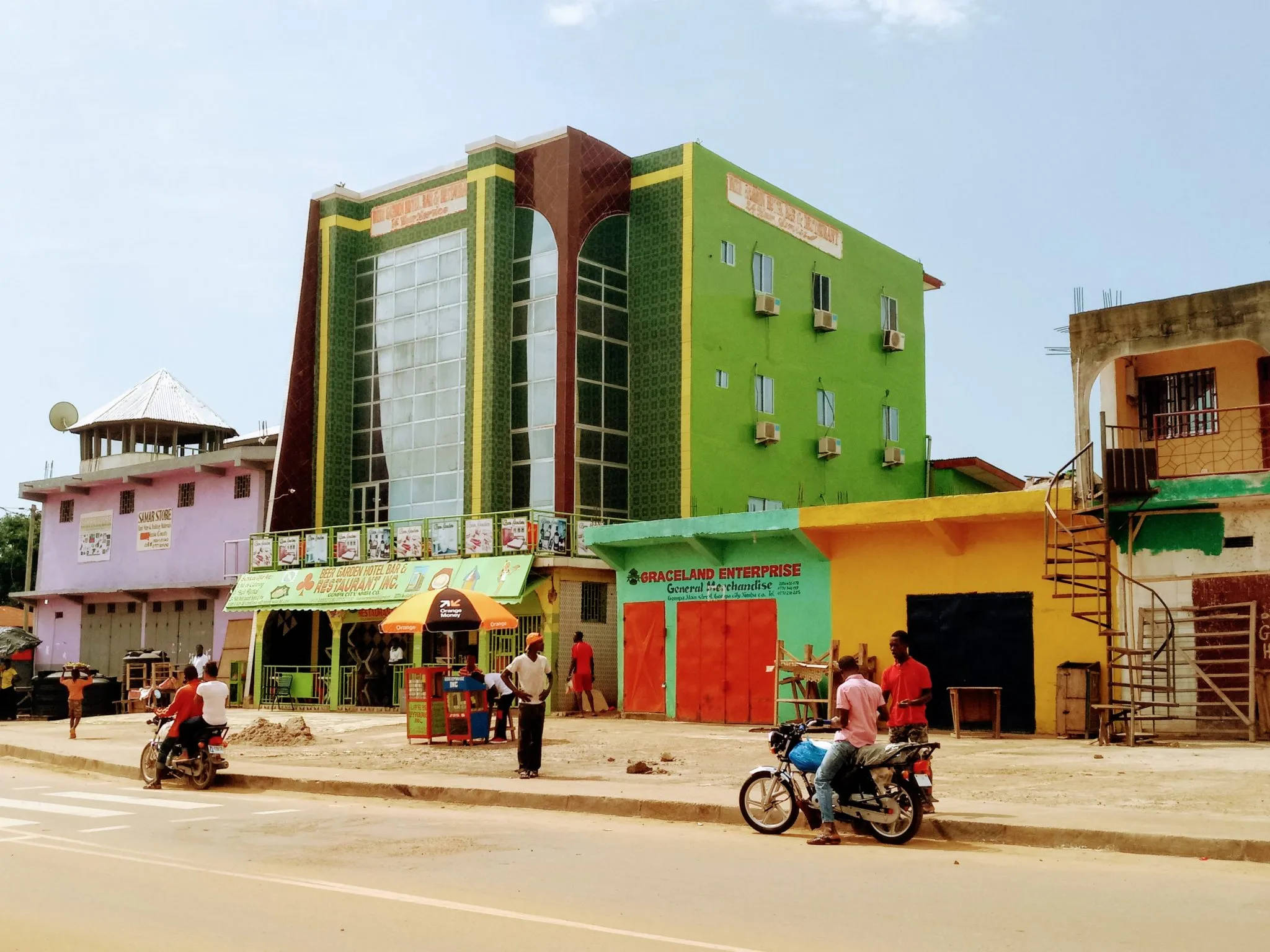 Liberia 桌布 壁紙 — 充滿活力的綠色和黃色玻璃幕牆建築，在繁忙的街道場景中高聳，摩托車和行人穿梭於晴朗的藍天下。
