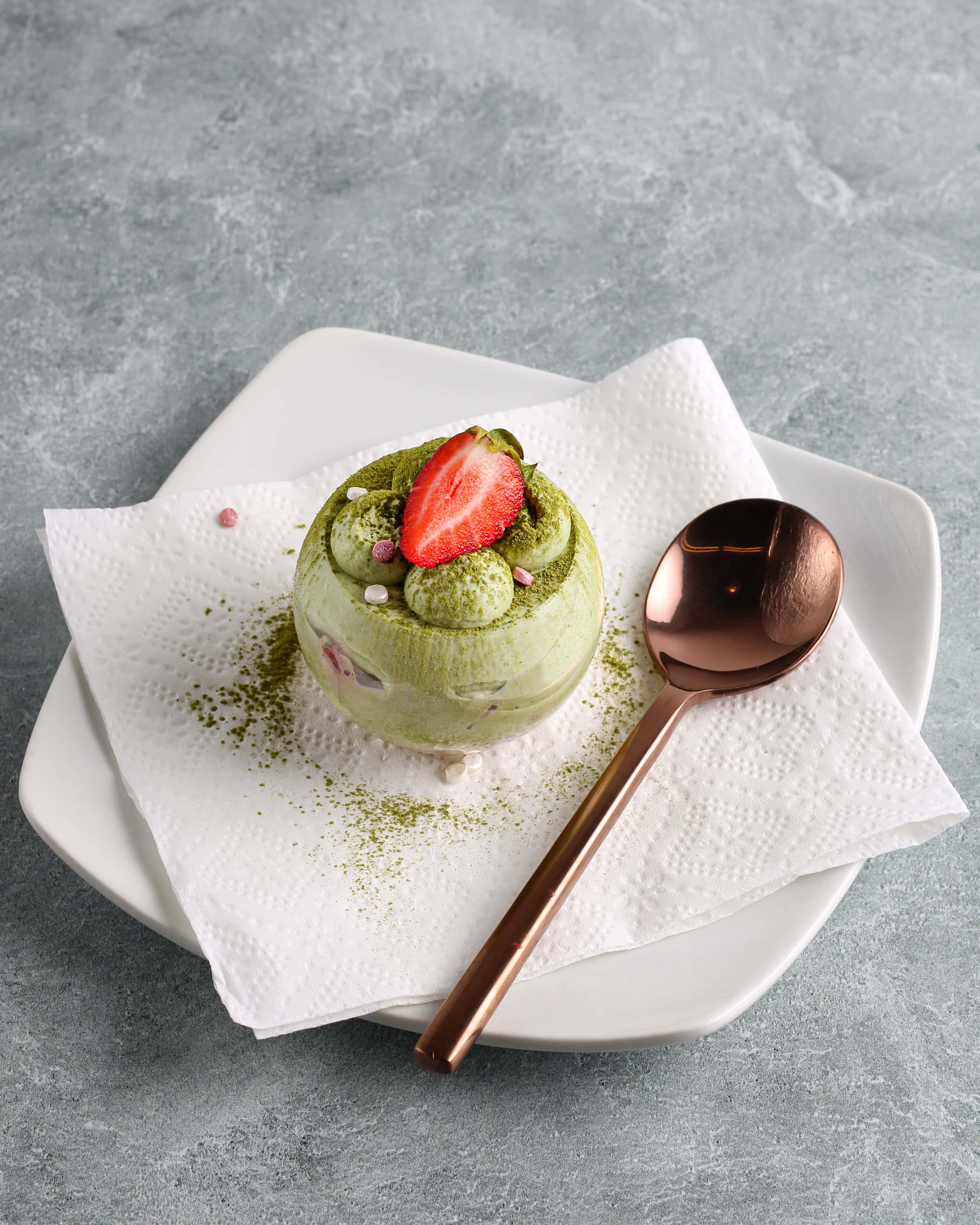 Japanese Sweets 桌布 壁紙 — 充滿活力的綠色抹茶甜品，上面放上新鮮草莓和粉紅色糖粒，優雅地放在白色碟子上，配上一把玫瑰金匙羹。
