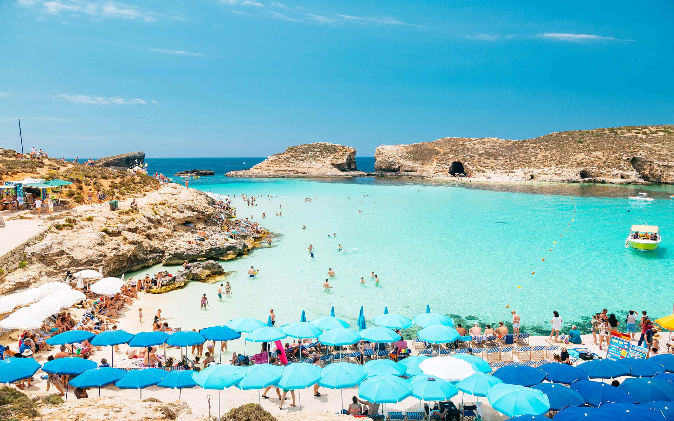 Malta 桌布 壁紙 — 充滿活力的藍綠色海水拍打著陽光普照的海灘，沙灘上擠滿了藍色遮陽傘和游泳者，天空清澈湛藍。