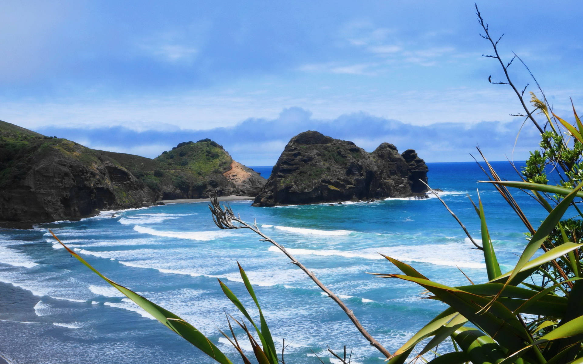 Beach Desktop 桌布 壁紙 — 充滿活力的藍綠色海浪拍打著崎嶇的懸崖，在明亮的藍天下，周圍環繞著綠色的海岸樹葉。