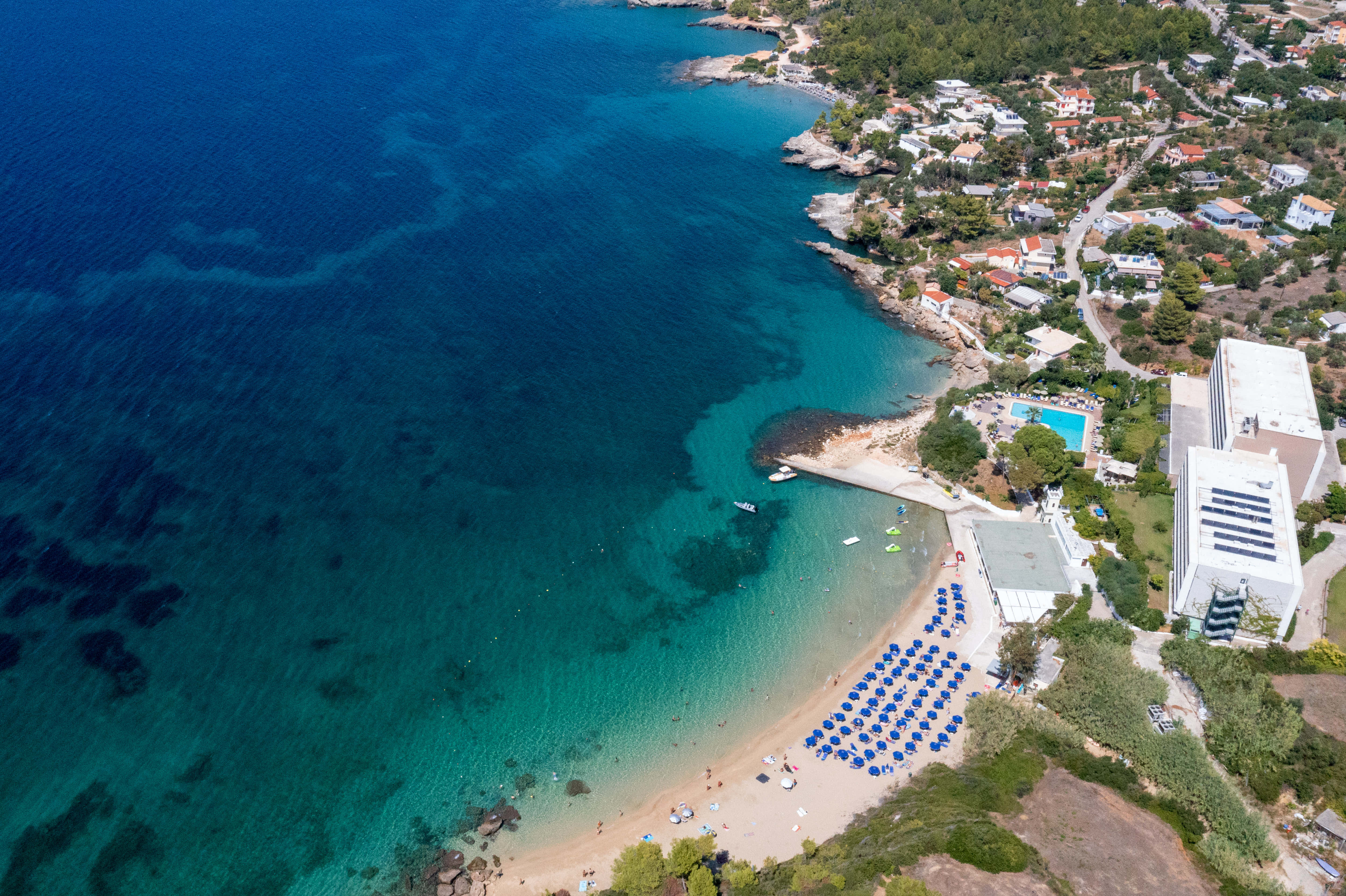 Mediterranean Beach 桌布 壁紙 — 充滿活力的藍綠色海灣鳥瞰圖，沙灘上排列著藍色遮陽傘，周圍環繞著沿海建築和綠植。