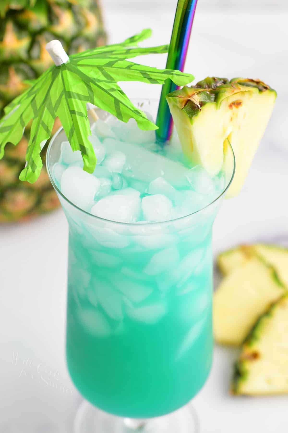 Blue Hawaii 桌布 壁紙 — 充滿活力的藍綠色熱帶飲料，裝滿冰塊，並以菠蘿片和綠色紙傘裝飾。