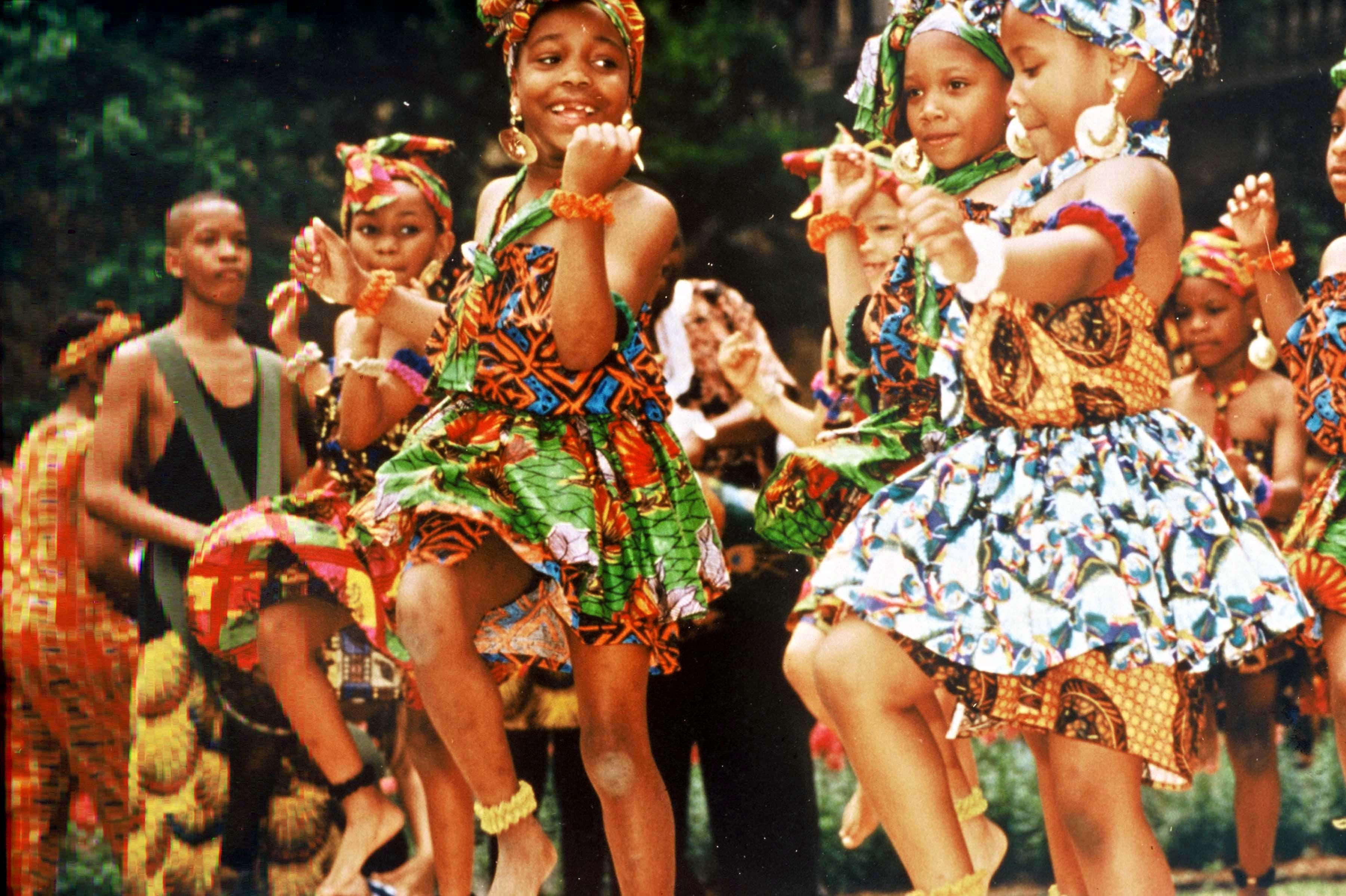 African Culture 桌布 壁紙 — 充滿活力的非洲兒童穿著色彩繽紛的圖案連衣裙，在明亮的日光下快樂地戶外跳舞。